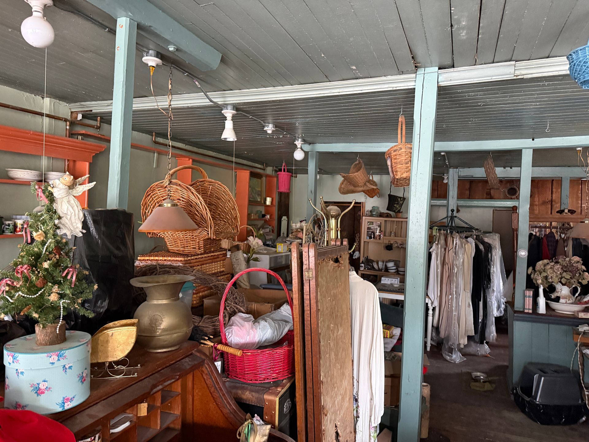 A vintage shop filled with hanging wicker baskets, clothing racks, antiques, and a small Christmas tree.