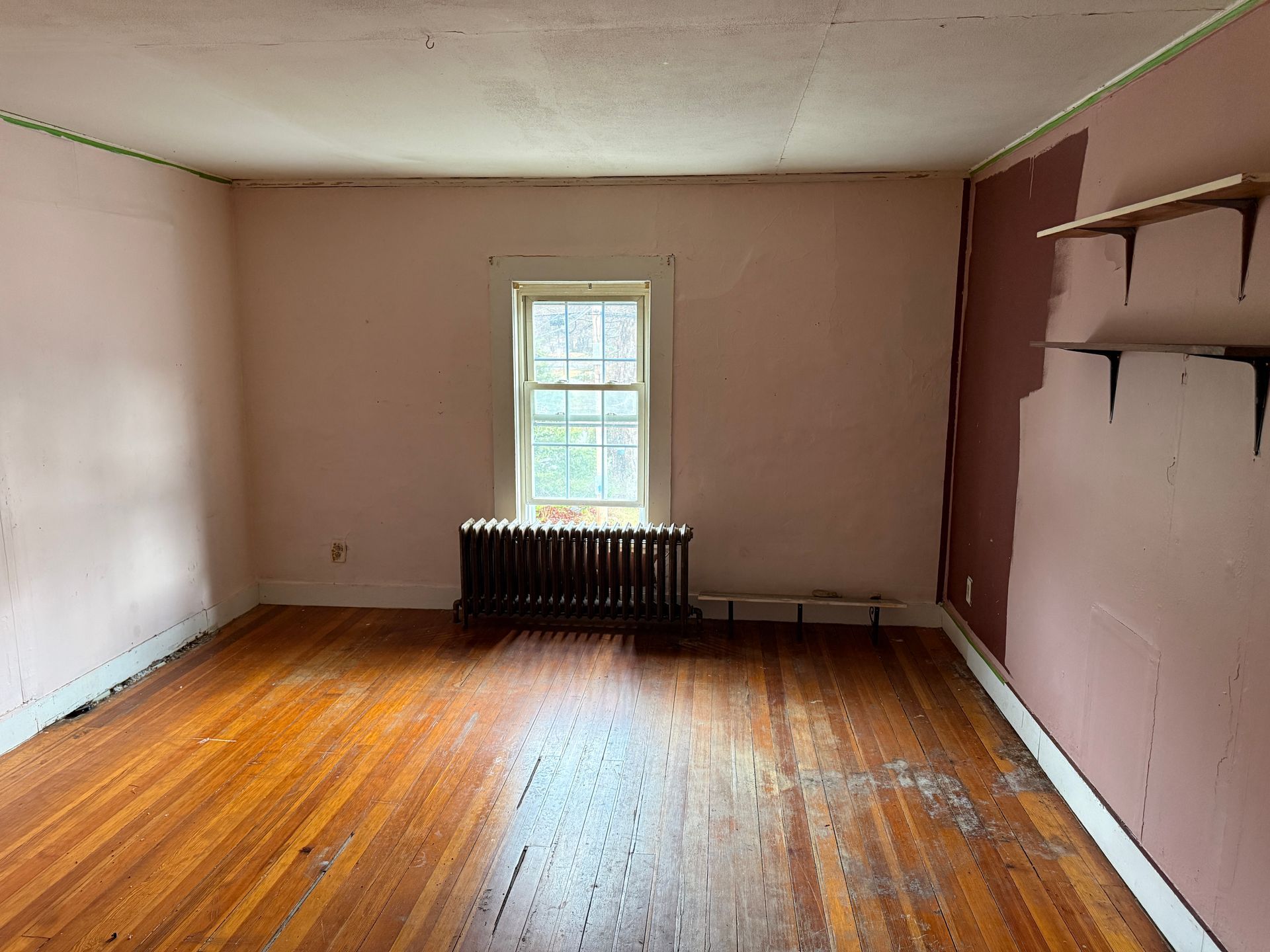 An empty room with pale pink walls, a center window, a dark radiator, polished wood floors, and two wall-mounted shelves.