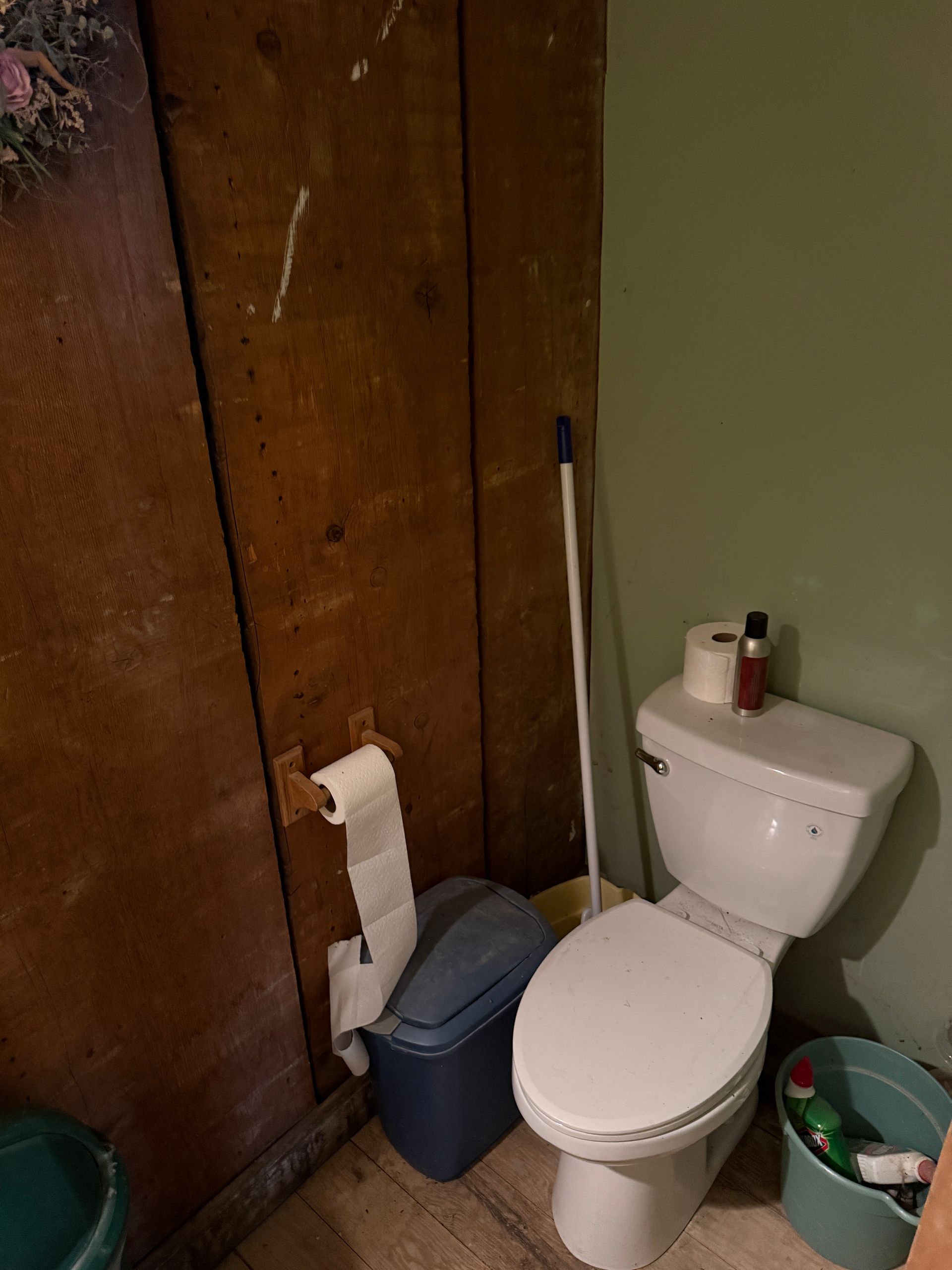 A bathroom corner with a white toilet, toilet paper holder on wood-paneled walls, a blue trash bin, and cleaning supplies.