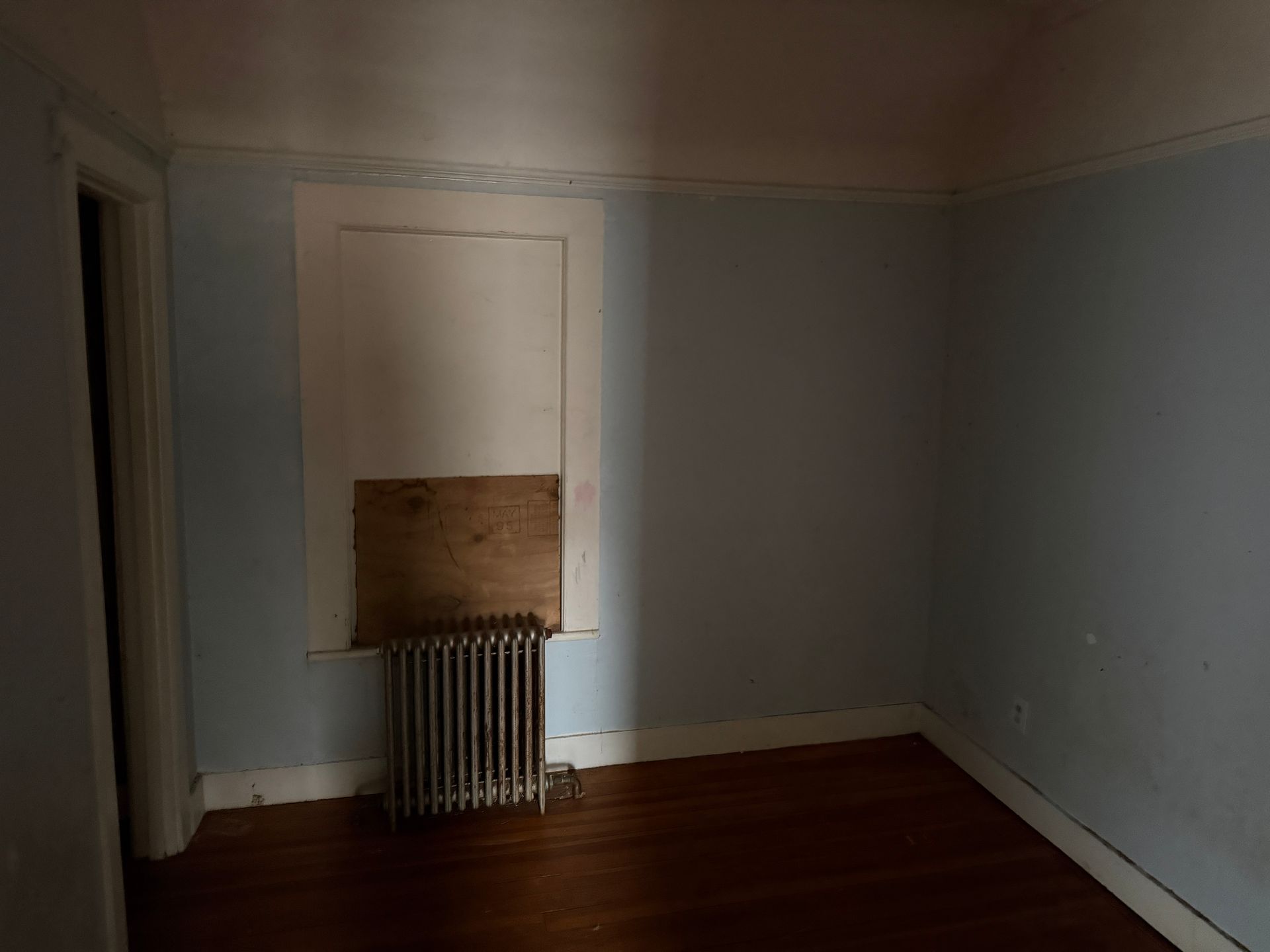 A corner of a room with light blue walls, a dark wood floor, a cast-iron radiator, and a window frame.