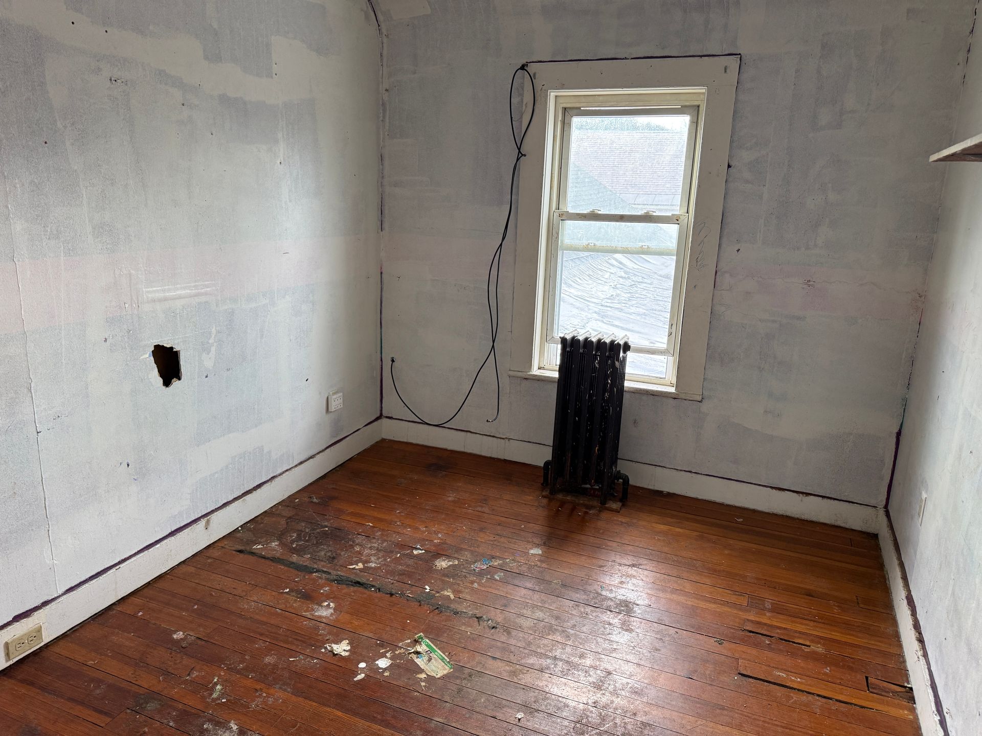 An empty room with wood floors, bare walls showing seams, a window, and a black cast-iron radiator.