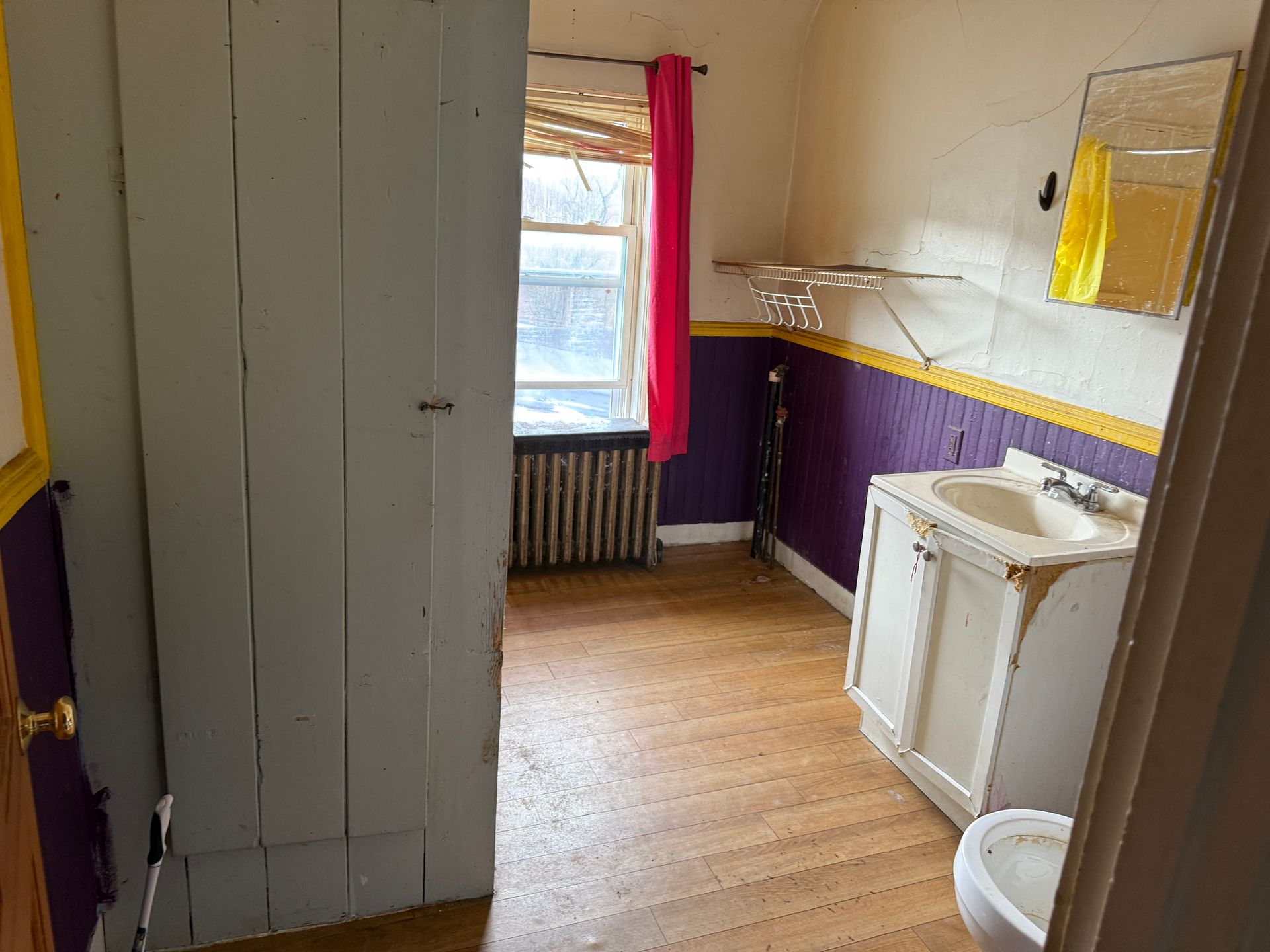 A bathroom with a white vanity, a toilet, a radiator near a window with pink curtains, and purple and yellow wall trim.