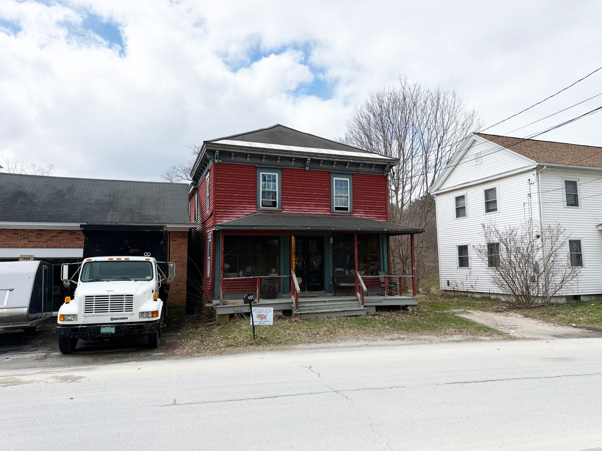 A ed two-story house between a brick building with a white truck and a white wooden house along a road.