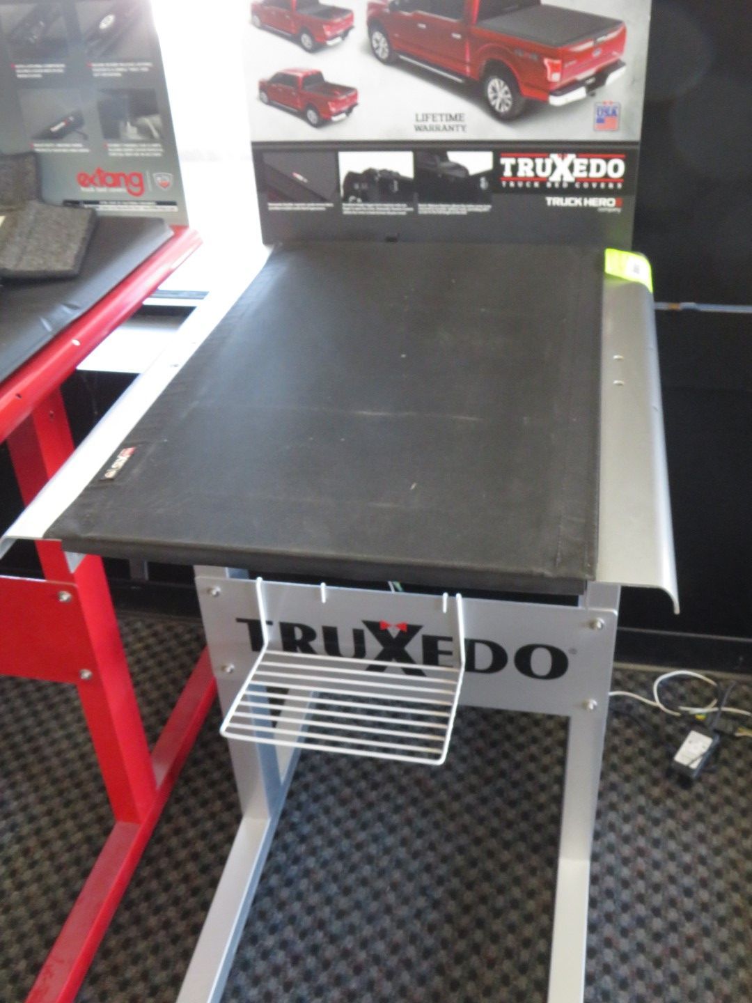A display stand for Truxedo truck bed covers, featuring a black cover on a metal stand with a wire basket.