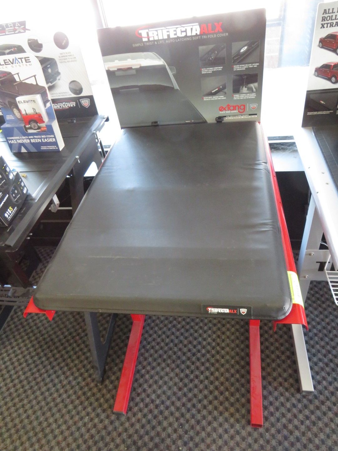 Black truck bed cover on a red and silver stand in a store. Product branding visible on the box and cover.
