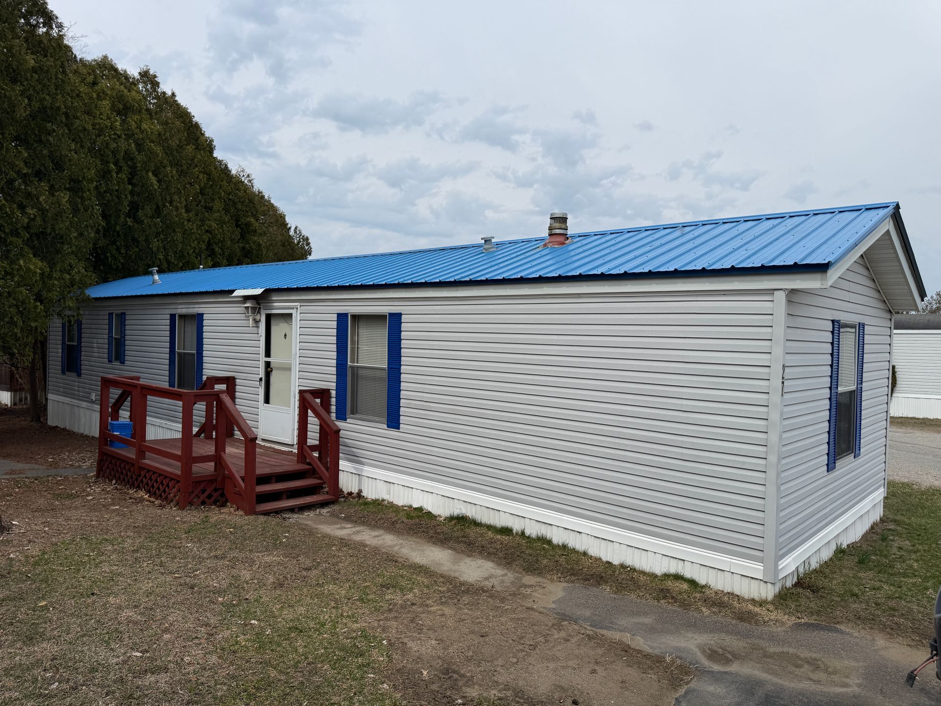 A light gray mobile home with a bright blue metal roof, blue window shutters, and a small red wooden deck entrance.