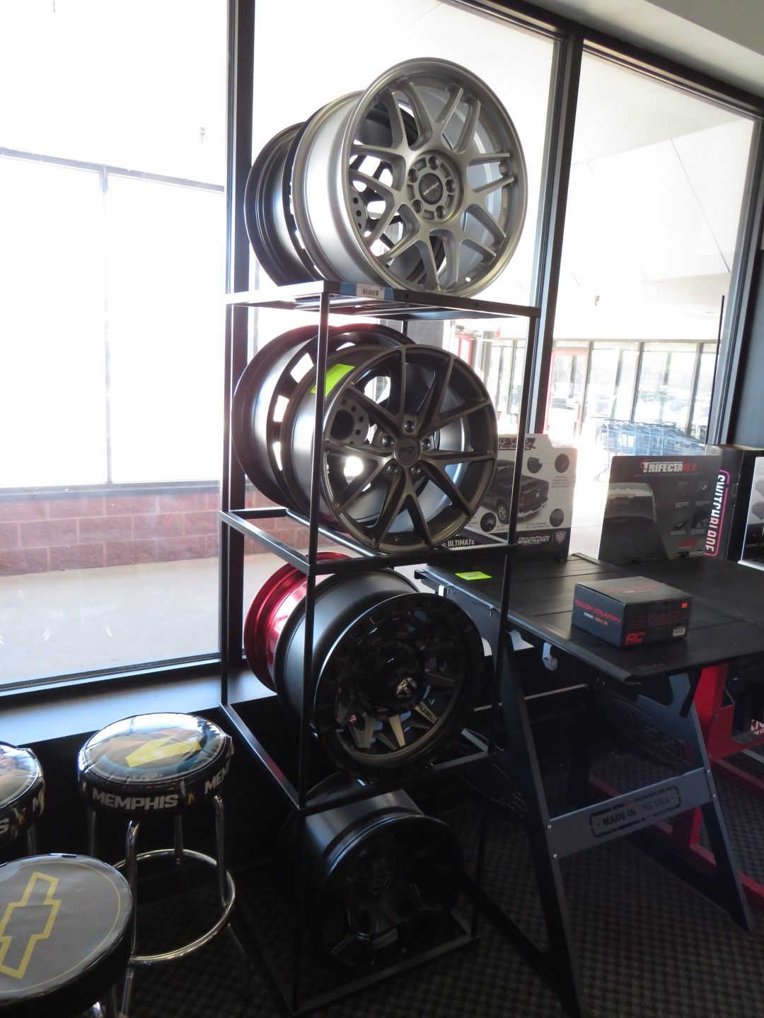 Wheels on display rack in showroom, various colors: silver, black, red. Window view, stools.