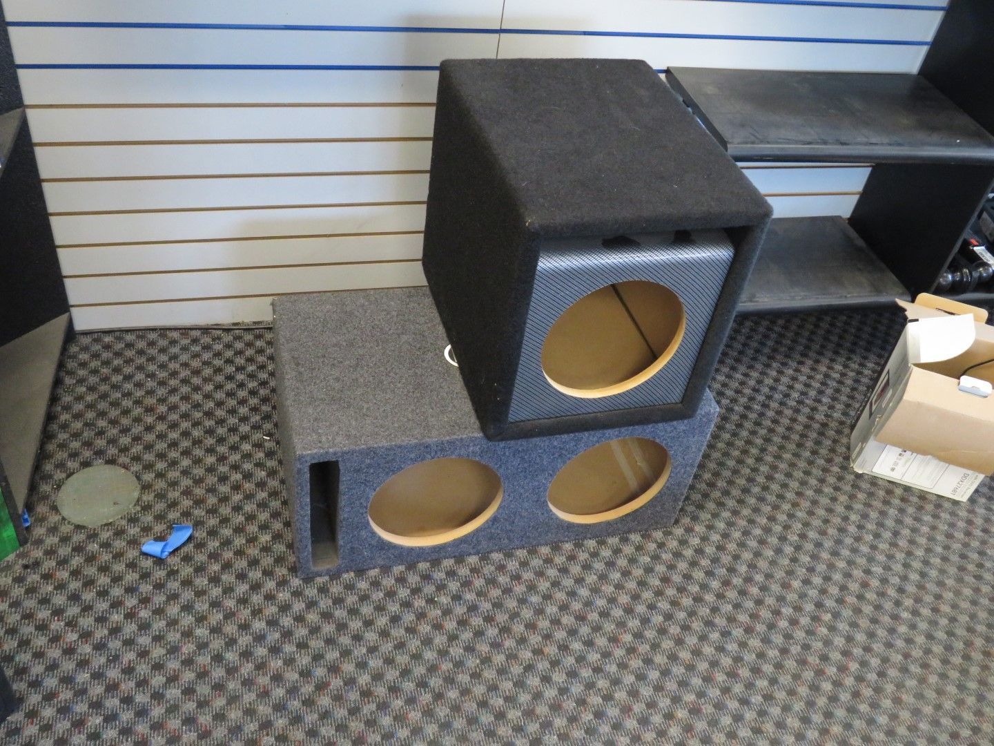 Two gray subwoofer boxes stacked on patterned carpet in a shop setting.