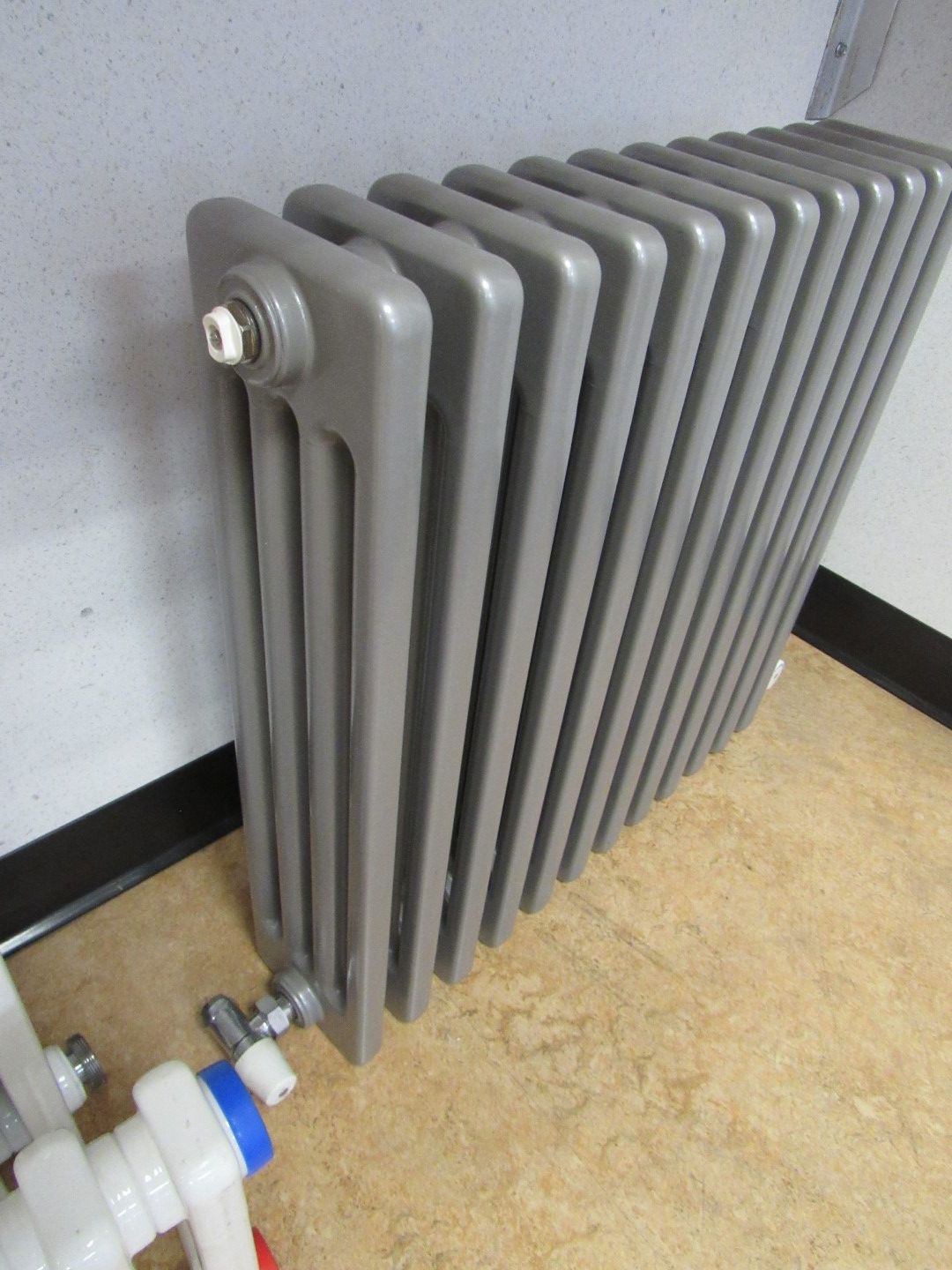 Gray radiator against a wall, connected to pipes on a beige floor.