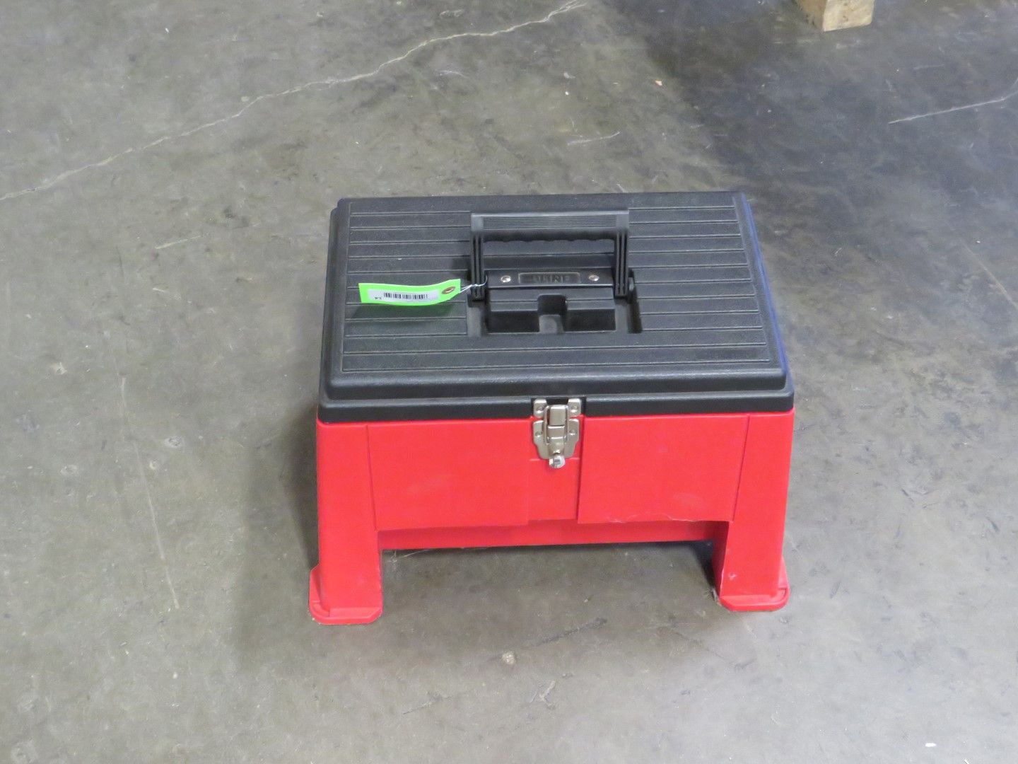 Red and black tool box on a gray concrete floor, with a handle on the black lid and latch on the red body.