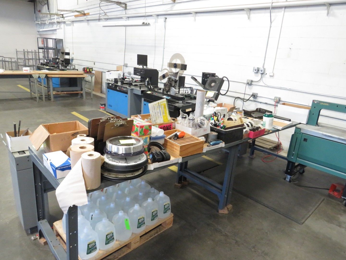 A workshop area with machinery, tools, and supplies; multiple workbenches and shelving units are visible.