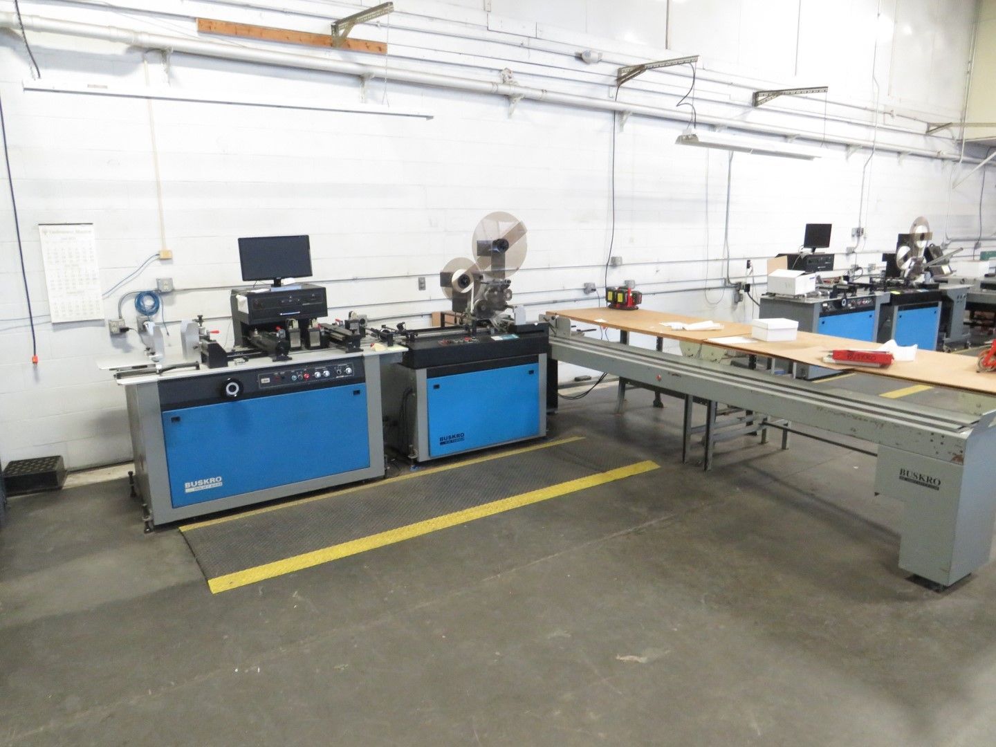 Blue industrial machines in a factory with a long wooden table and yellow safety line.