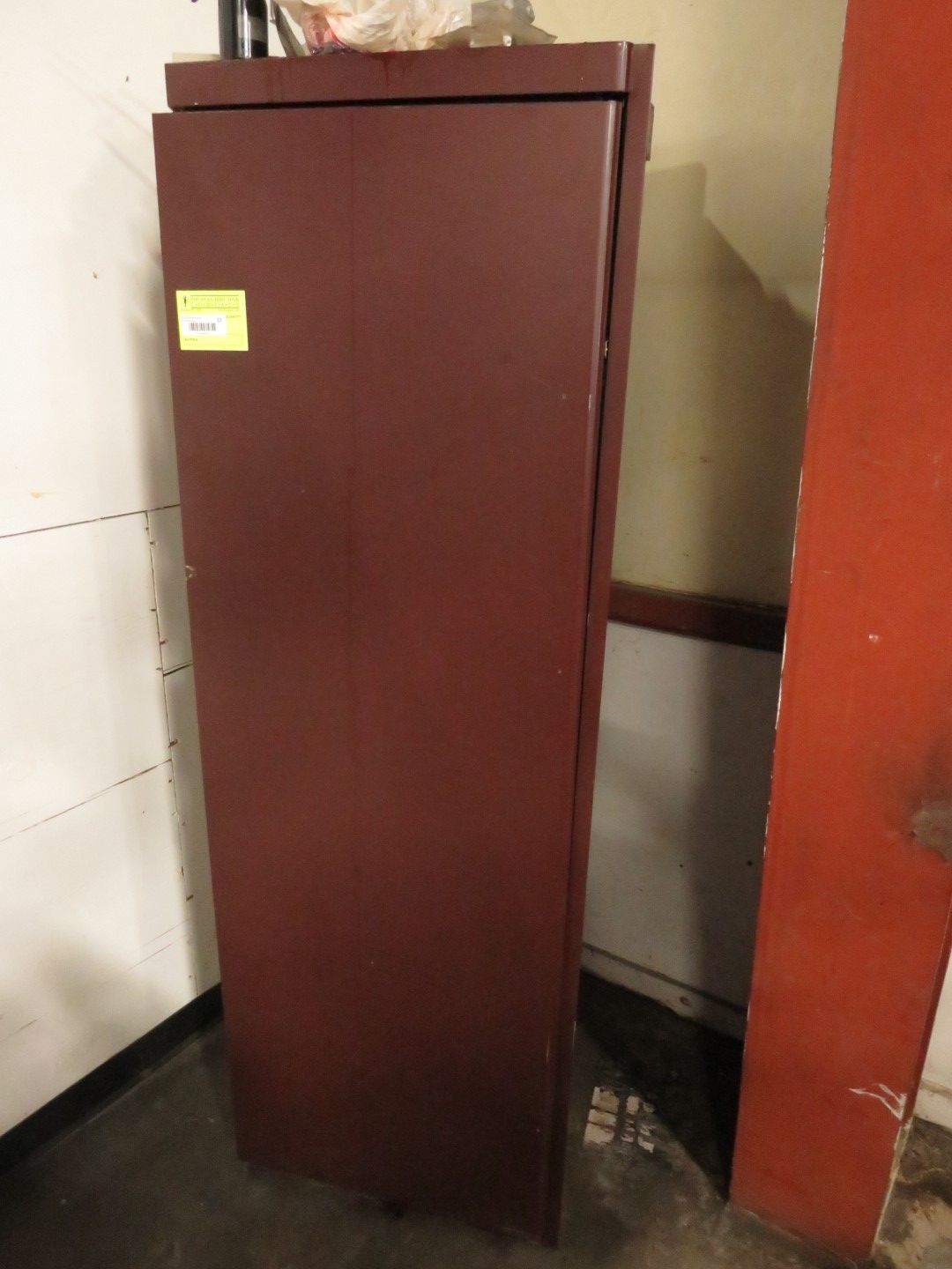 Tall, maroon metal cabinet with a closed door, indoors. Yellow tag on the front.
