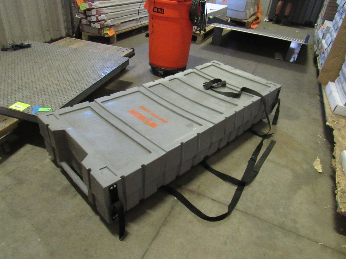 Gray rectangular storage case with black straps and orange text in a warehouse setting.