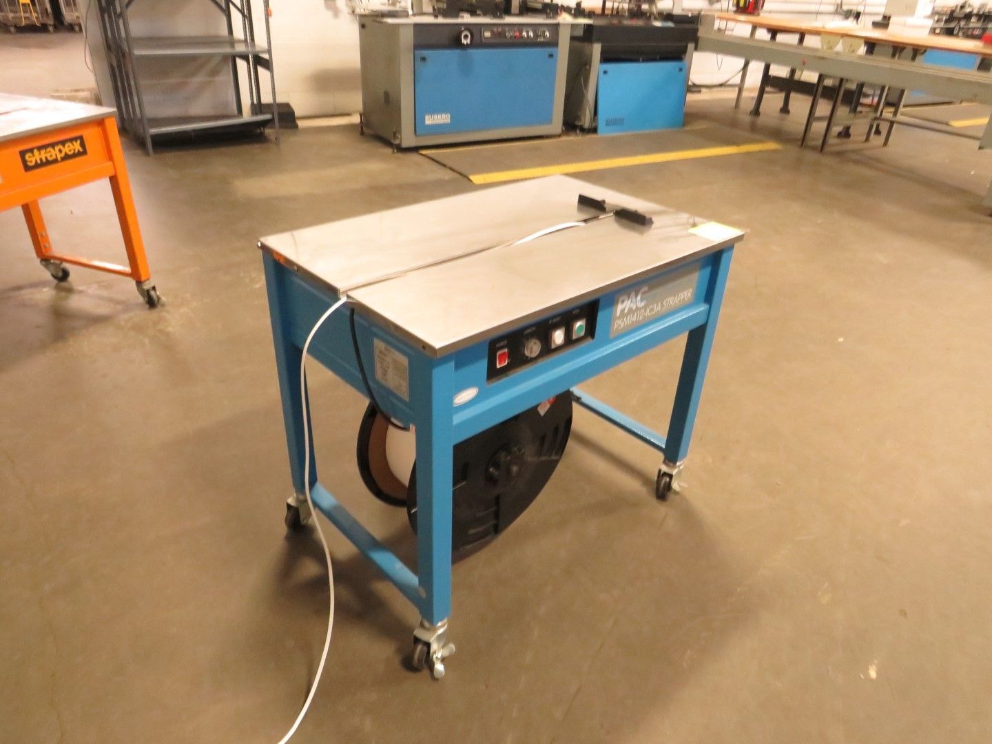 Blue industrial strapping machine on wheels, steel table, electrical panel.