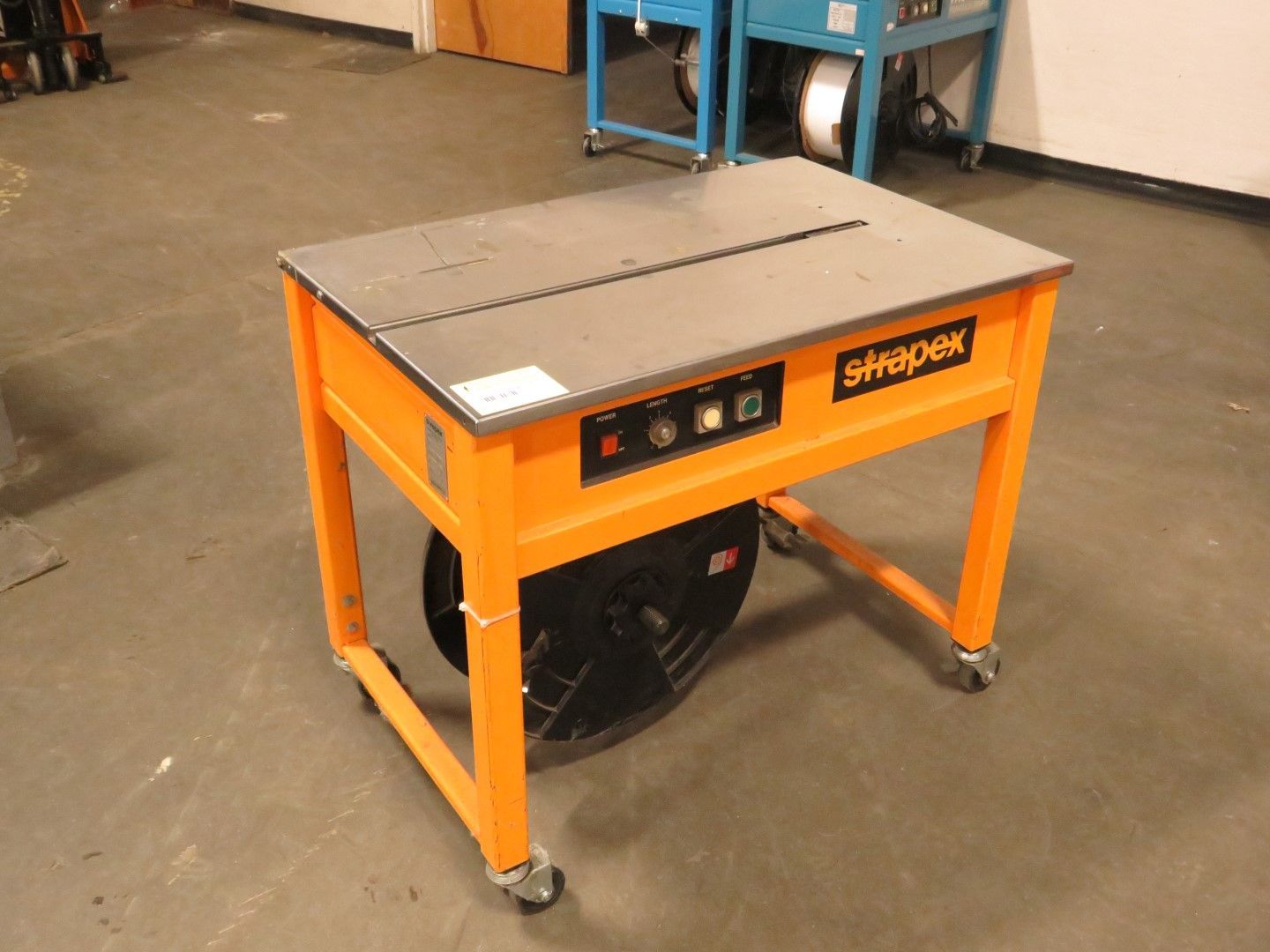 Orange and gray strapping machine on wheels. Shows controls and a spool of strapping material.
