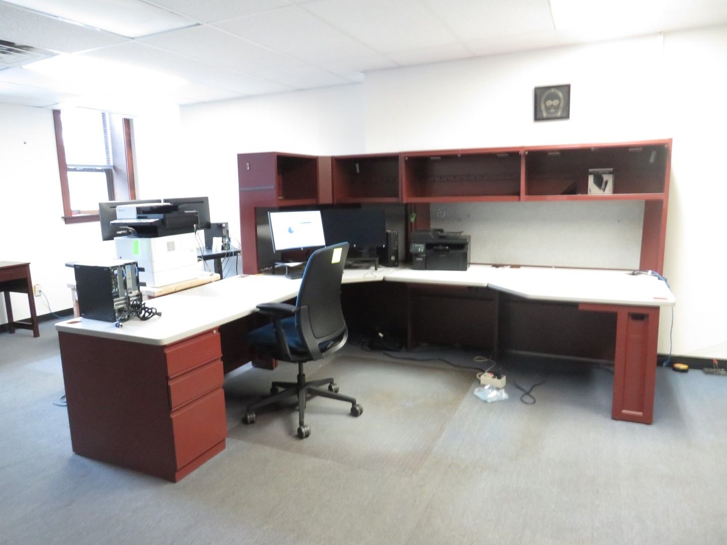 Office workspace: L-shaped desk with computers, printer, overhead storage, and rolling chair. Maroon and white colors.