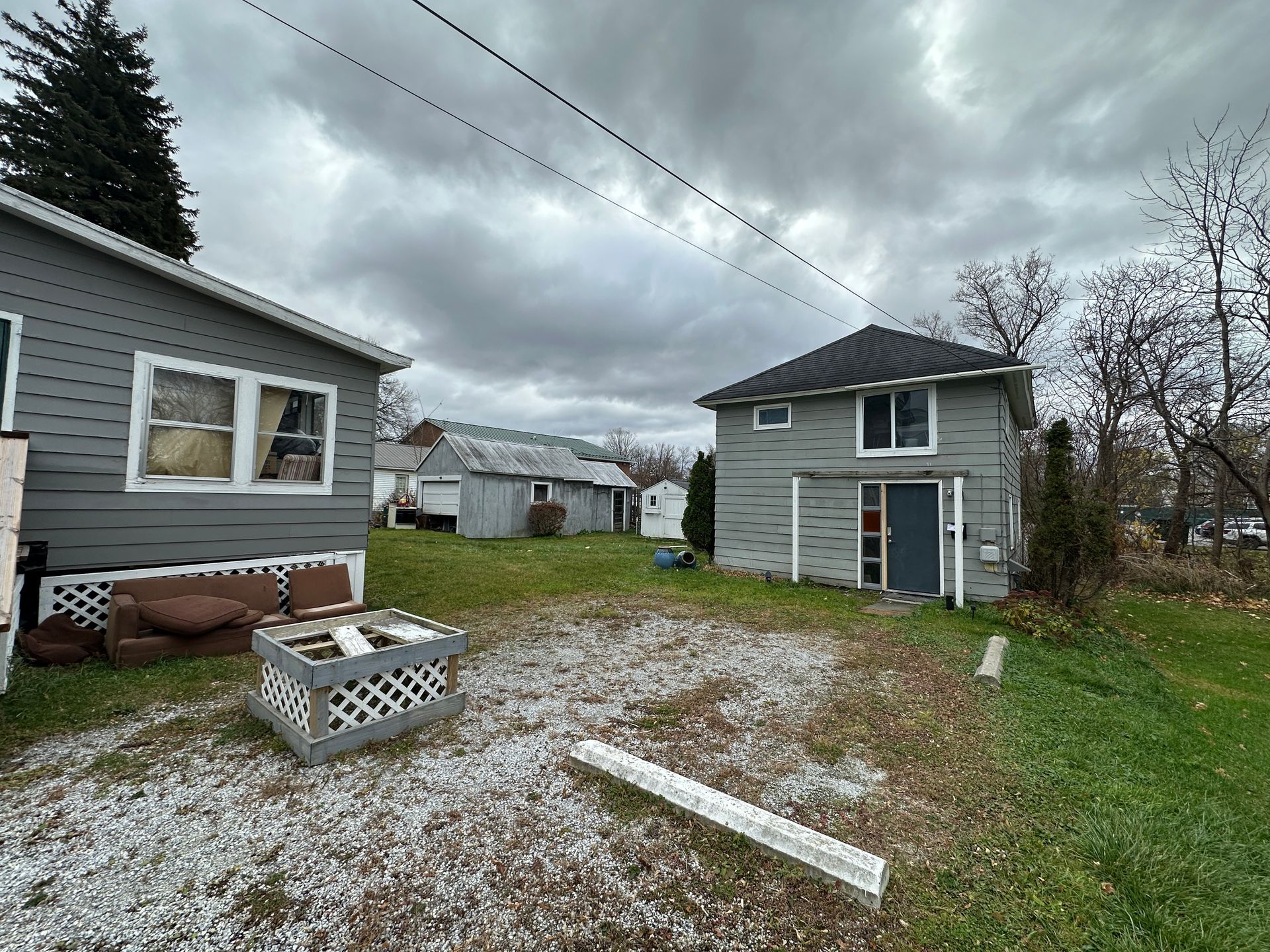 Two gray houses stand on a grassy lot under a cloudy sky, with a small shed and a gravel area in the foreground.