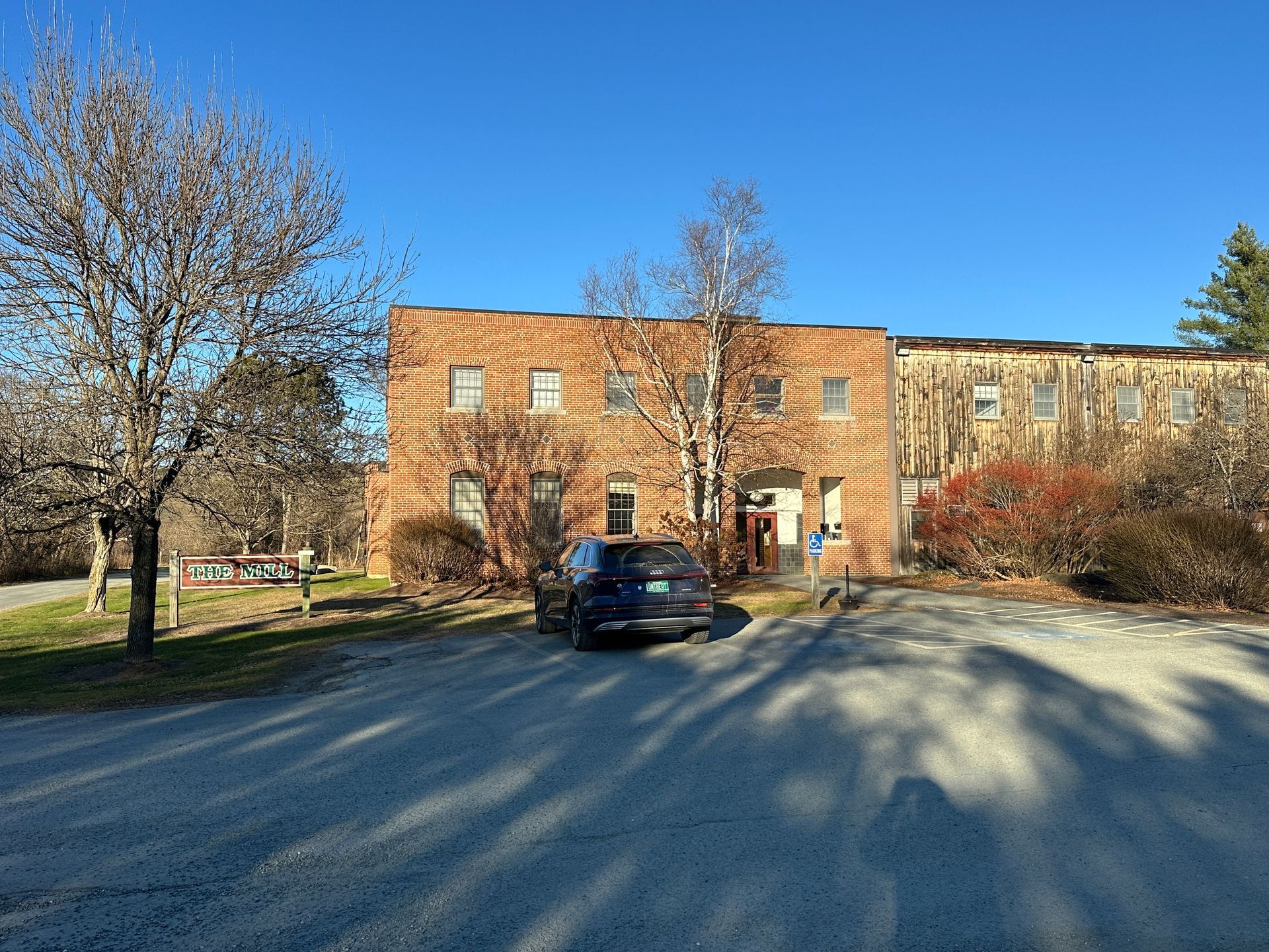 Brick building with a dark car parked in front; trees and bushes surround it on a sunny day.