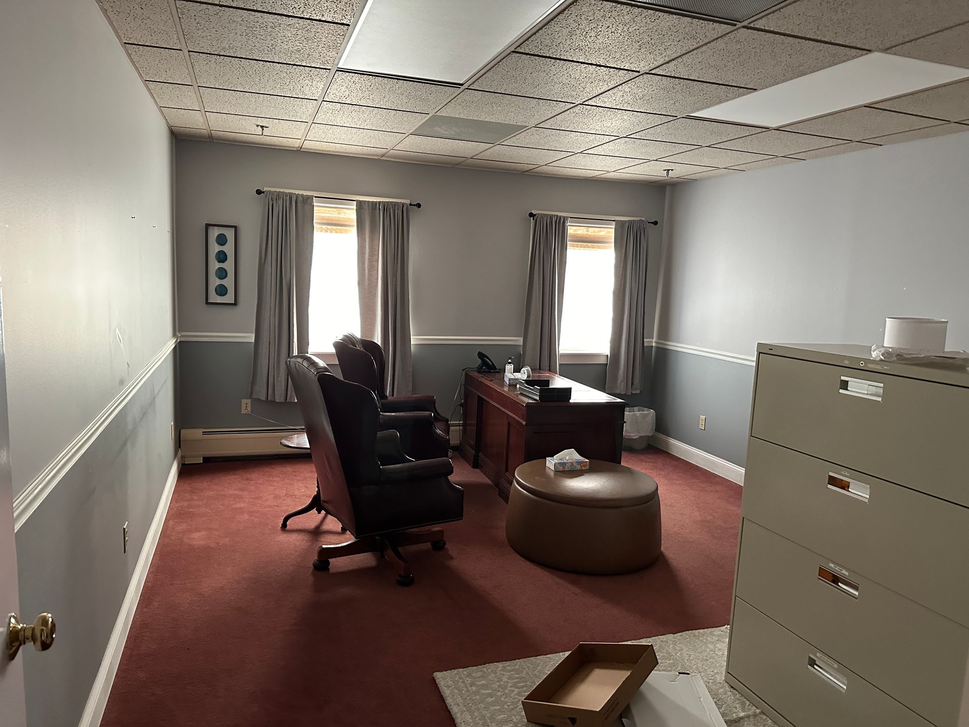 Empty office with gray walls, red carpet, desk, file cabinet, and two windows with curtains.