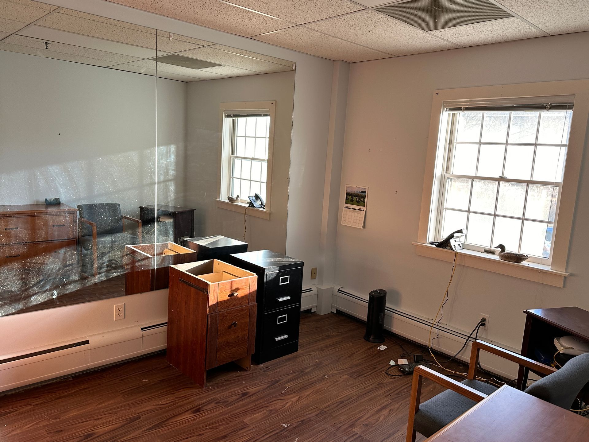 Empty office with two windows, a mirror, wooden floor, and filing cabinets.