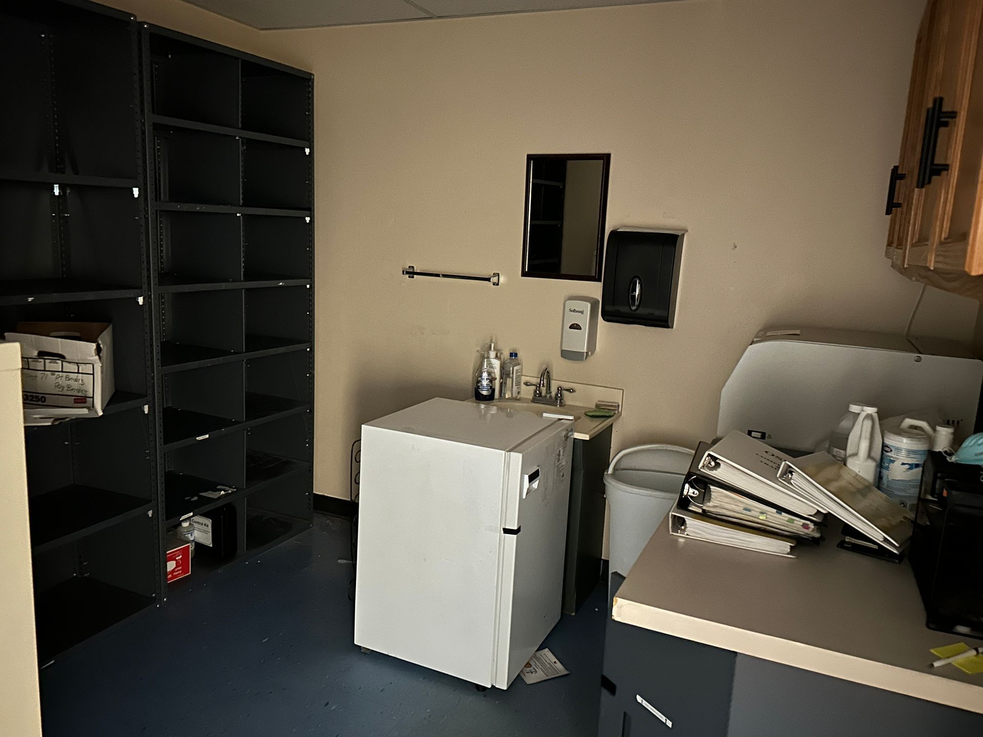A room with shelves, a refrigerator, sink, and cabinets; possibly a medical office or utility space.