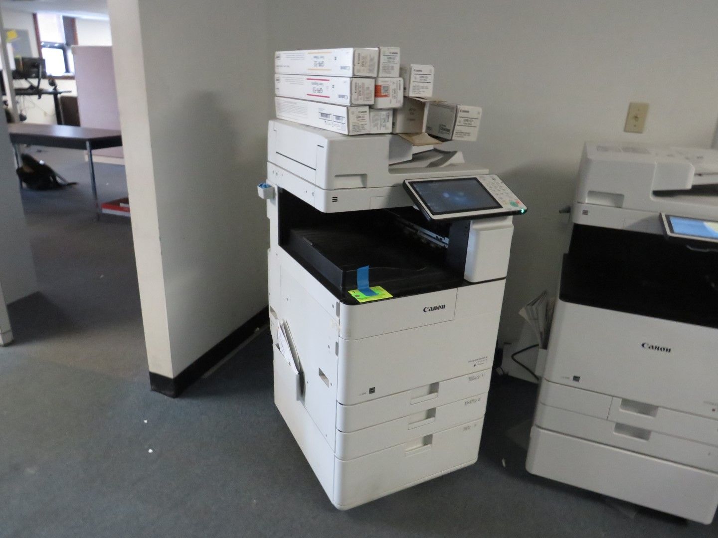 White office copier with stacked paper and another copier to the right.