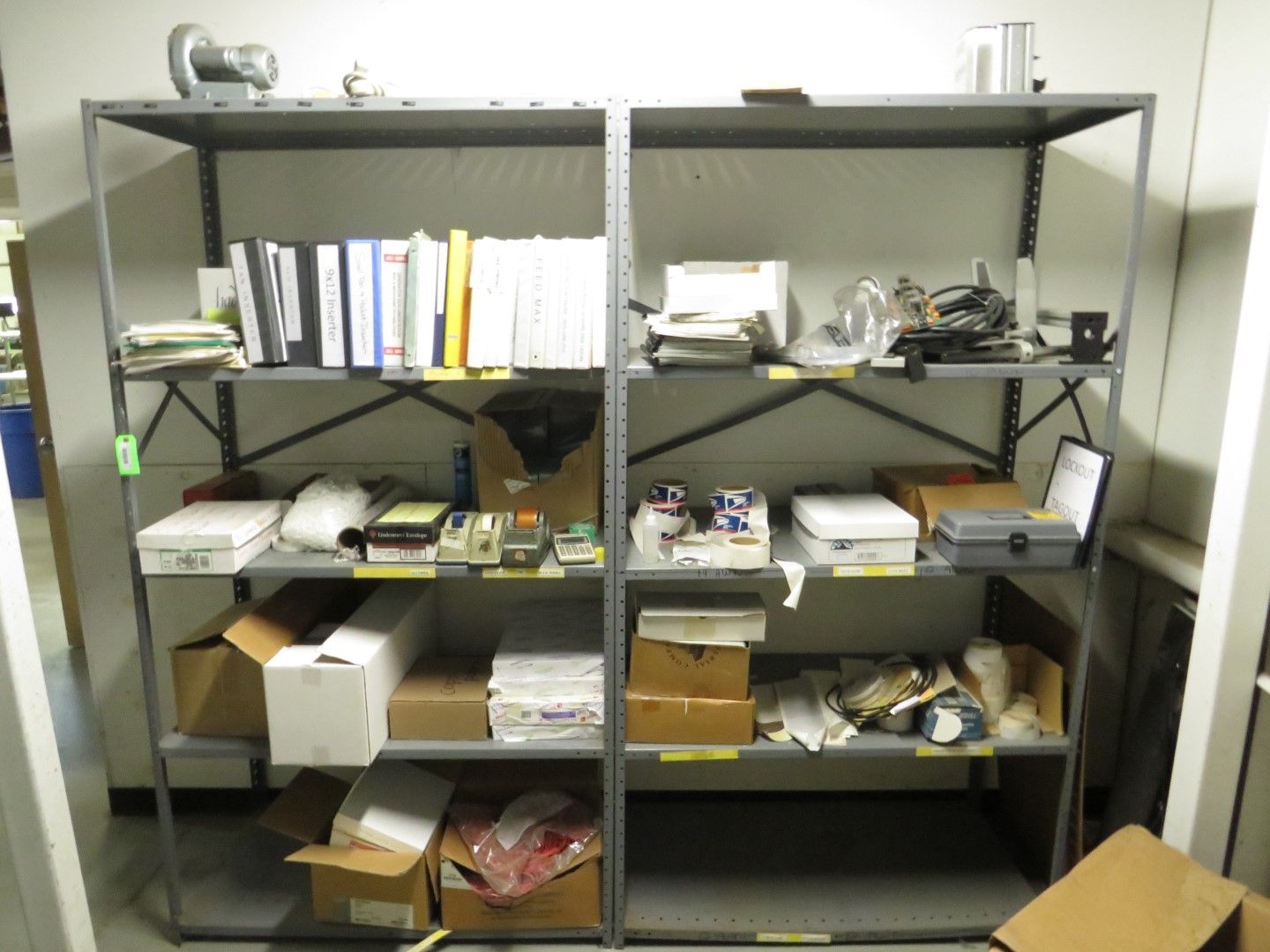 Metal shelving unit filled with various office supplies, binders, and boxes in a storage room.