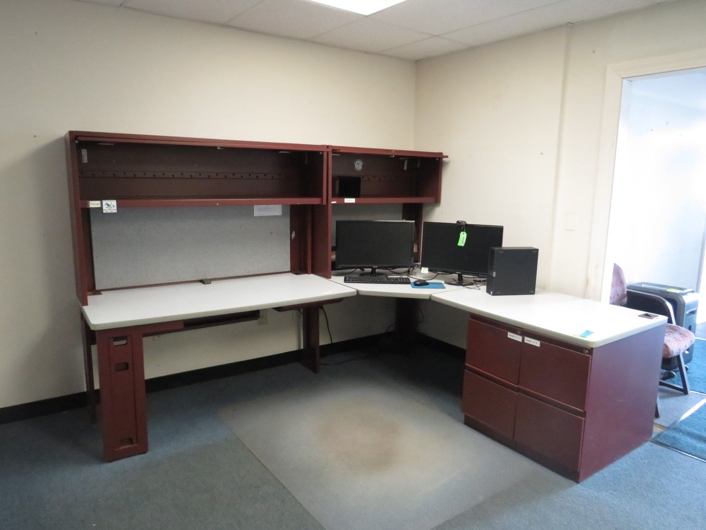 L-shaped office desk with overhead storage, two monitors, and a cabinet in a room.