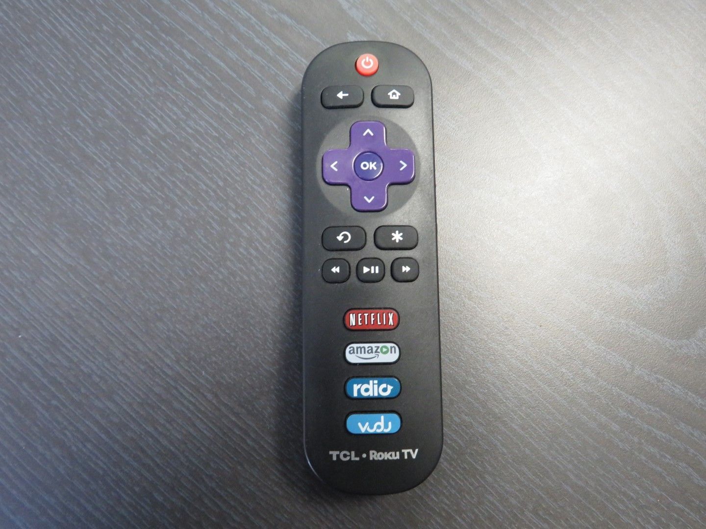 Black TCL-Roku TV remote on a gray surface, featuring directional buttons and shortcut keys.