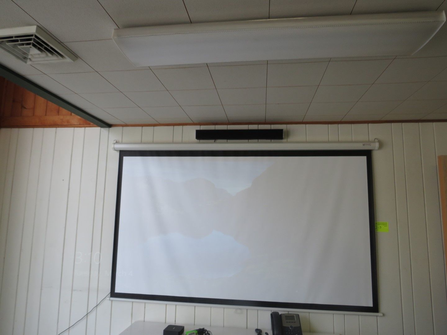 White projector screen mounted on a wall, with a speaker bar above it. A ceiling light is above the screen.