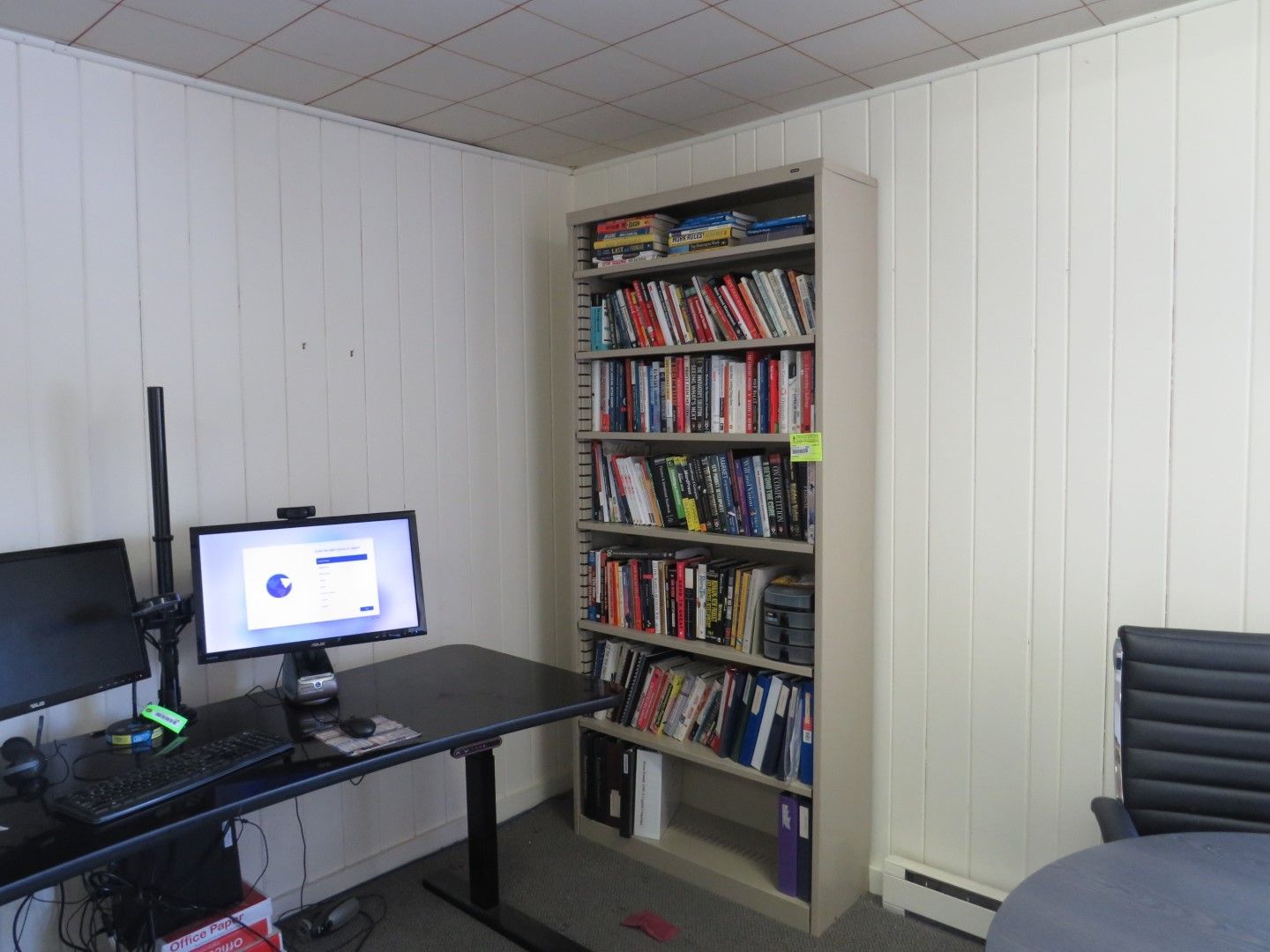A home office with a computer, desk, and bookshelf filled with books. White paneled walls.