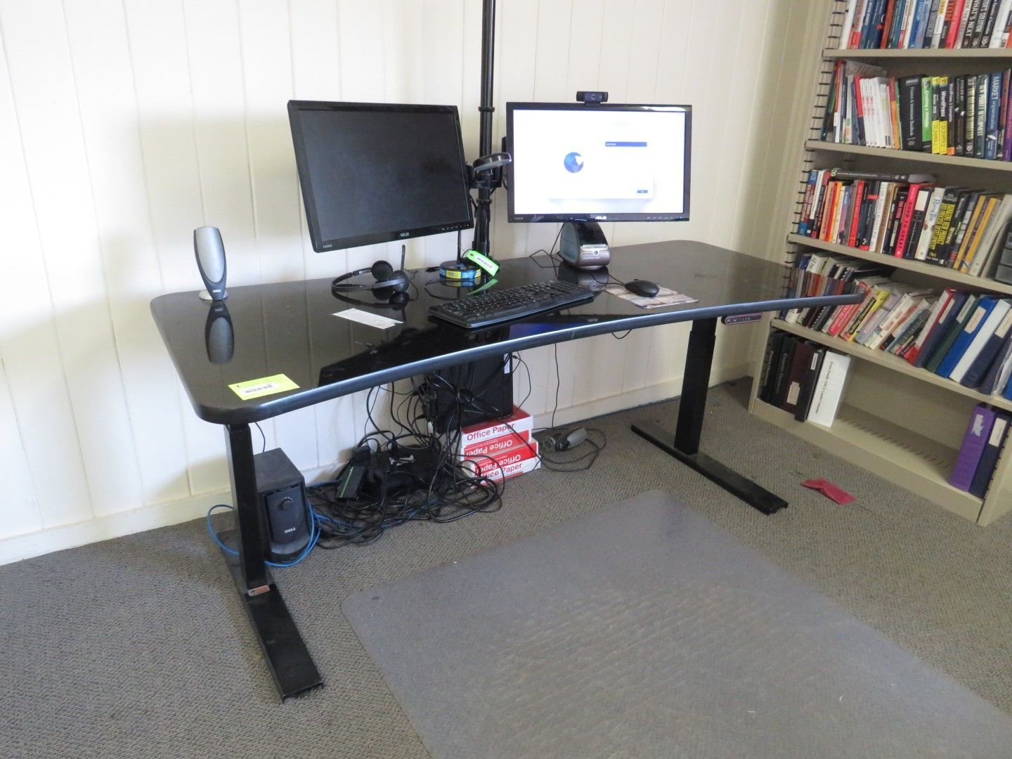 Black sit-stand desk with two monitors, keyboard, and cables against a white wall and bookshelf.