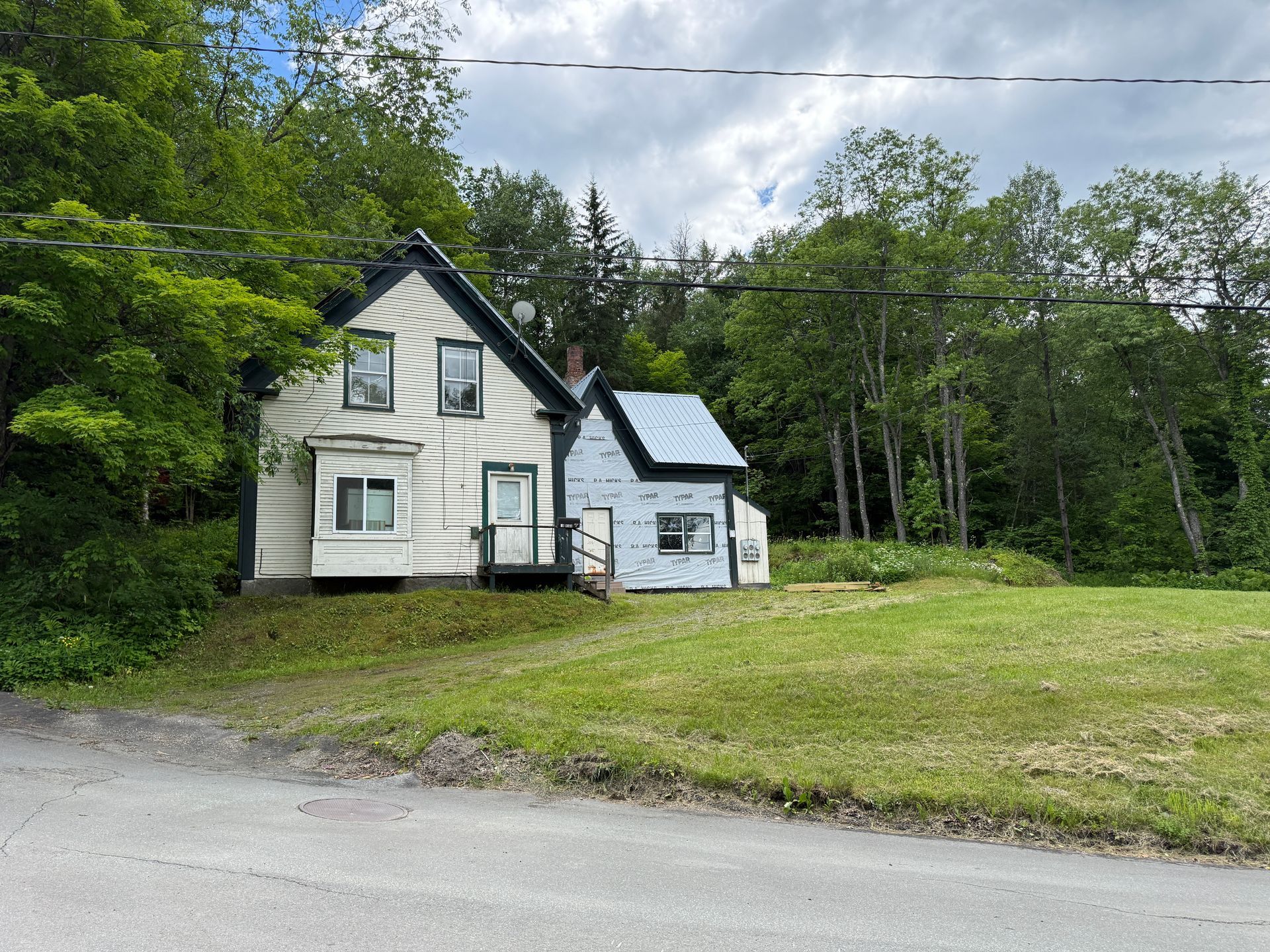Small white house with dark trim beside a wooded area and grassy yard under a cloudy sky