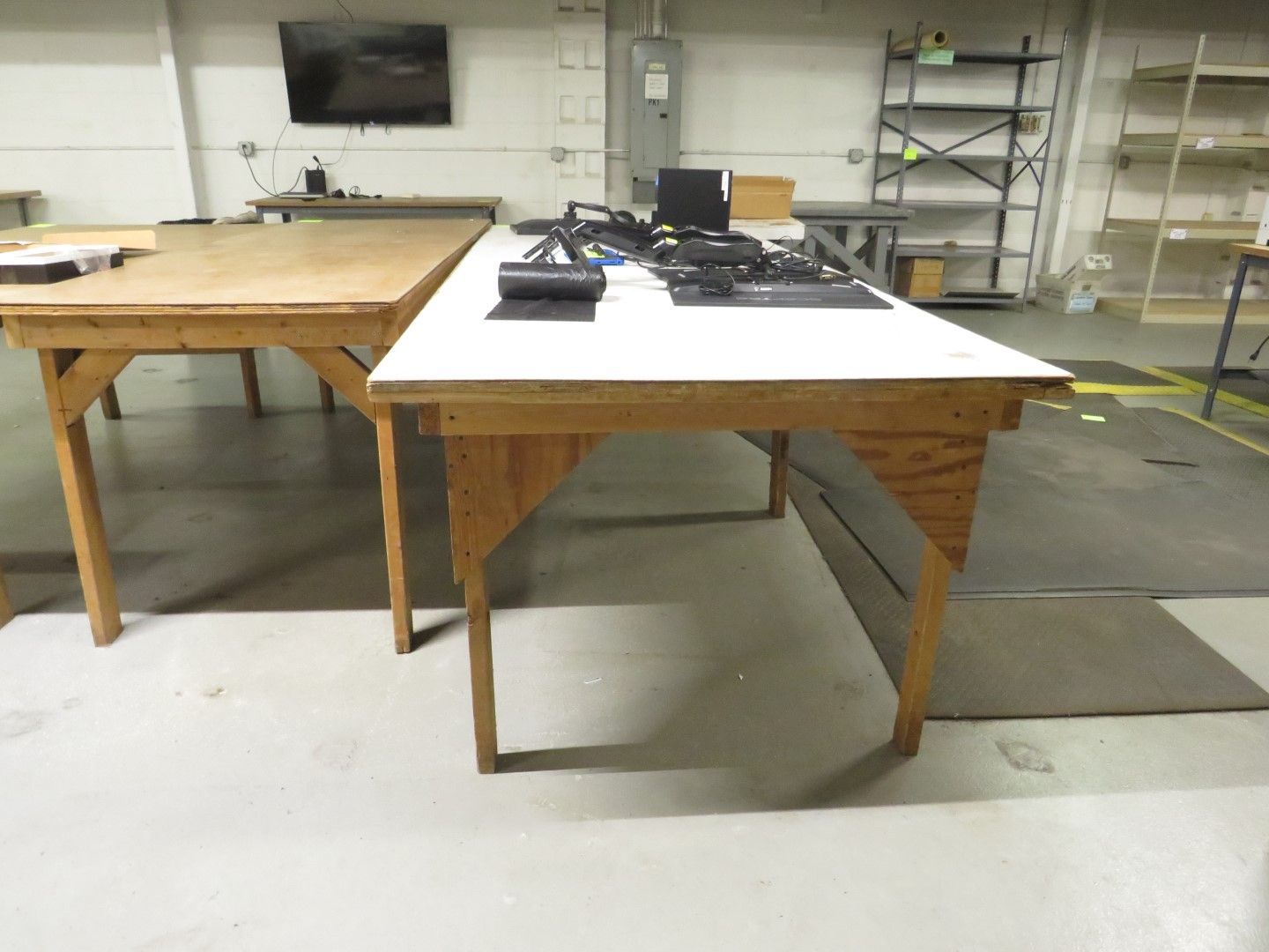 Two wooden work tables in a factory setting. One table is light wood, the other is white, with equipment on top.