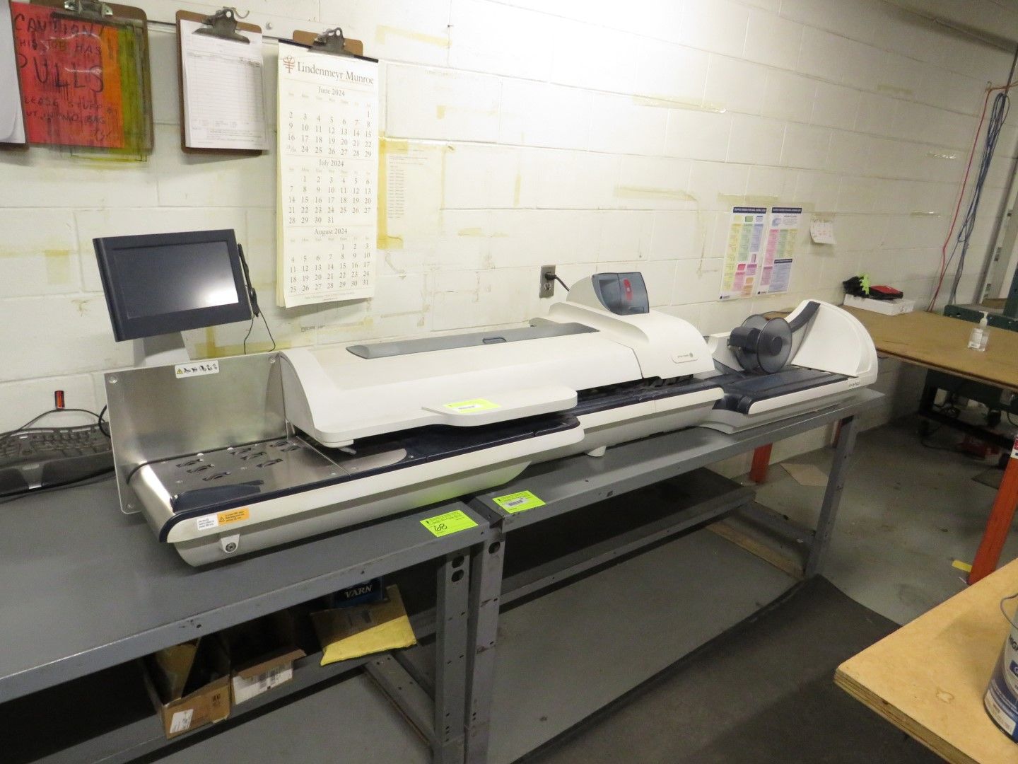 Mailing machine on a gray table in a warehouse setting, next to a computer monitor, and a wall calendar.