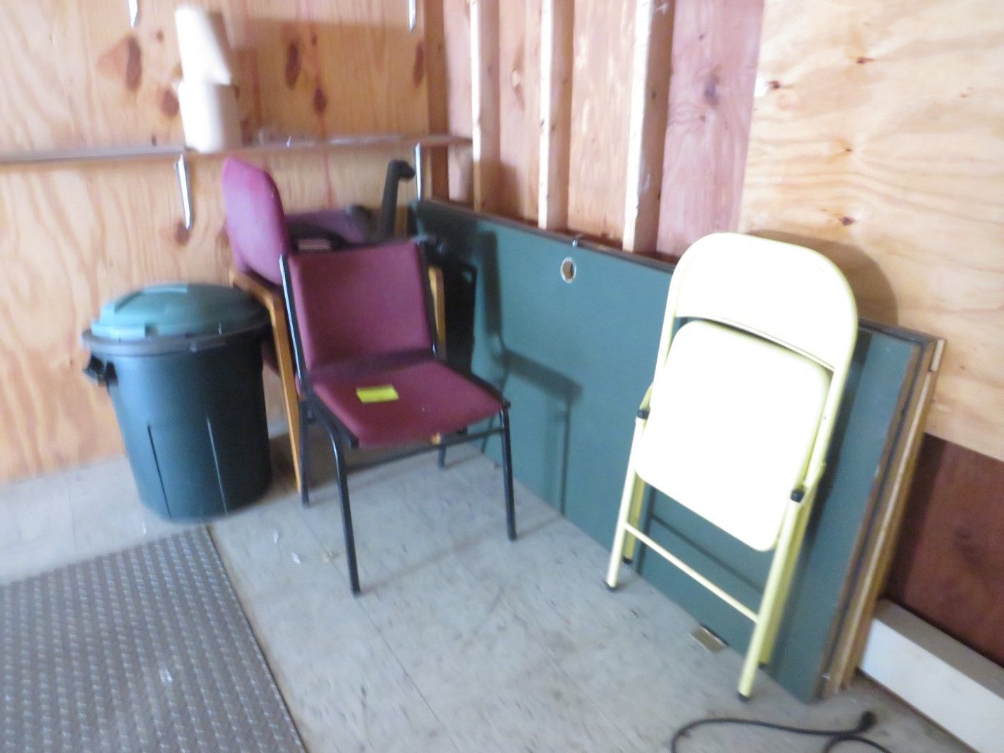 Storage space with a trash can, two chairs, a folding table, and a folded chair.