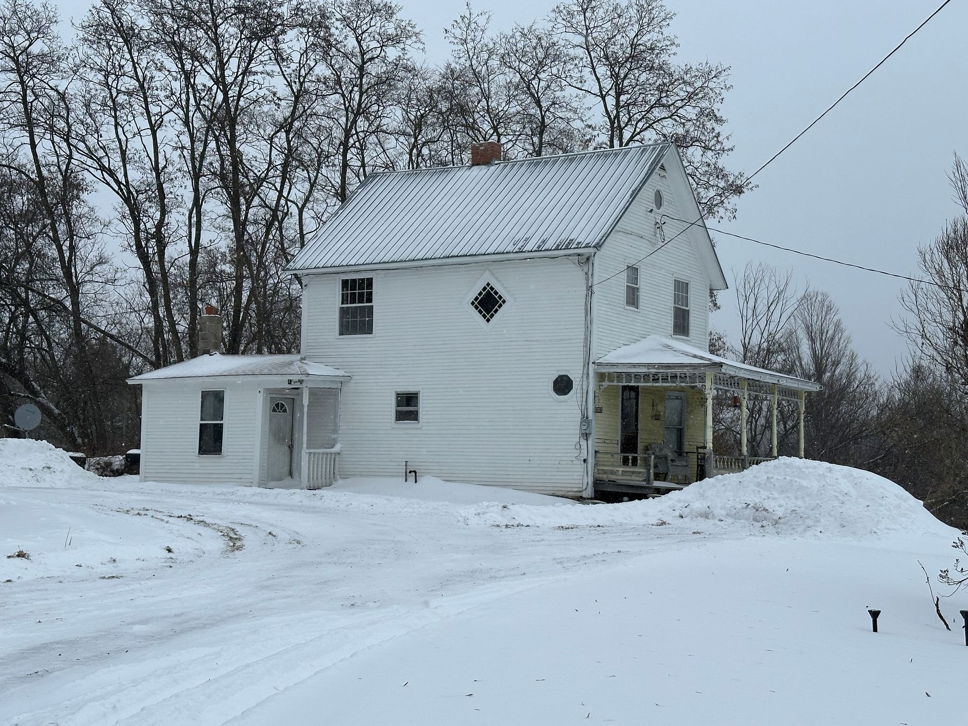 A two-story white farmhouse with a small addition sits in a snow-covered yard surrounded by bare trees under a gray sky.