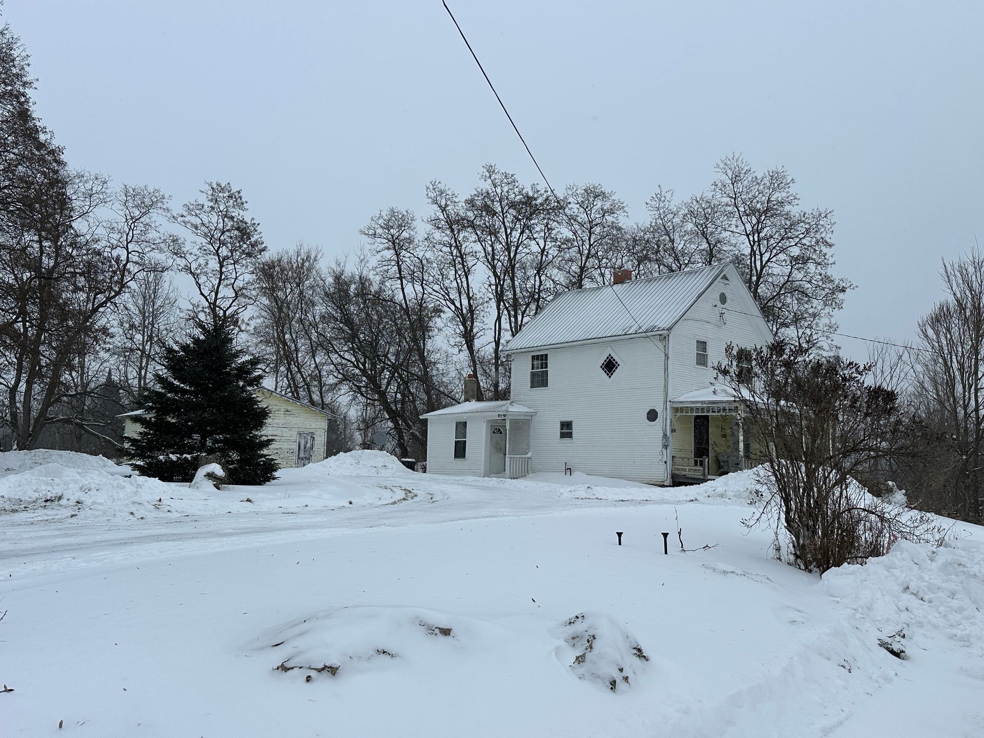 A white two-story house stands on a snow-covered yard on a cloudy winter day, with bare trees in the background.