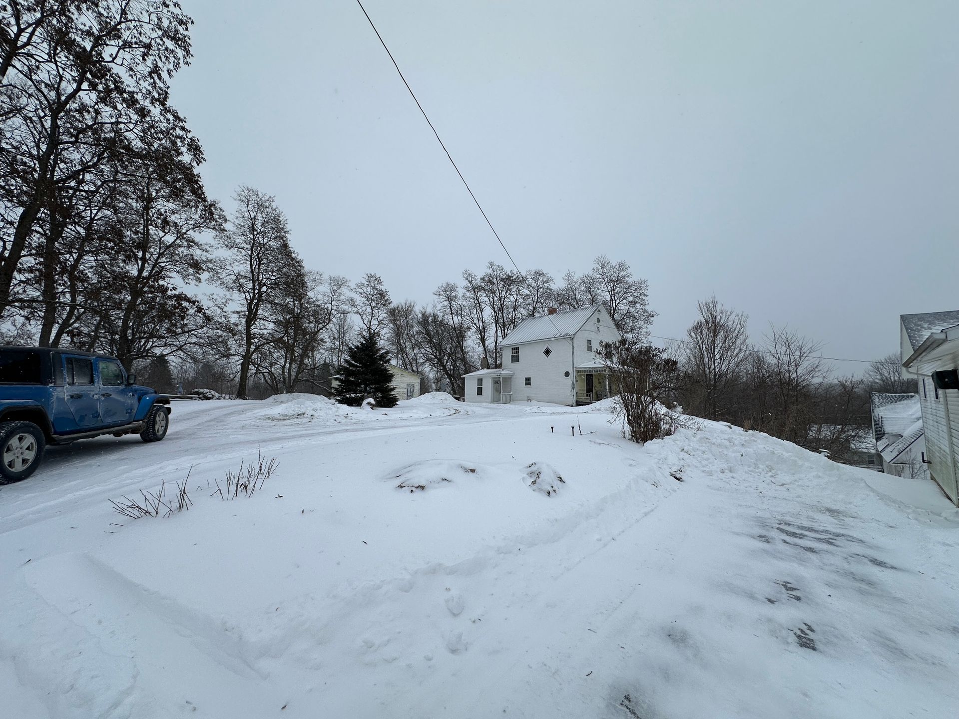 A blue truck parked on a snow-covered yard in front of a white house surrounded by trees on a cloudy winter day.