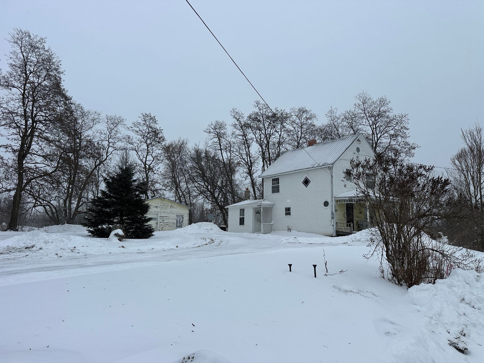 A white house and a smaller outbuilding stand in a snowy, wooded landscape under an overcast sky.