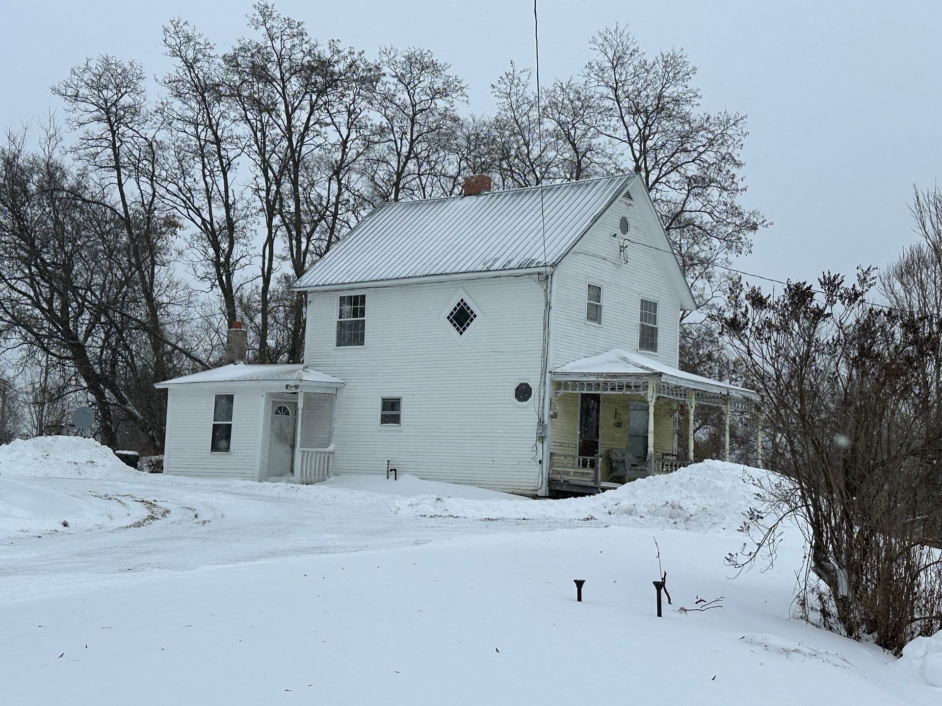 A white, two-story farmhouse stands surrounded by deep snow in winter with tall, bare trees in the background.