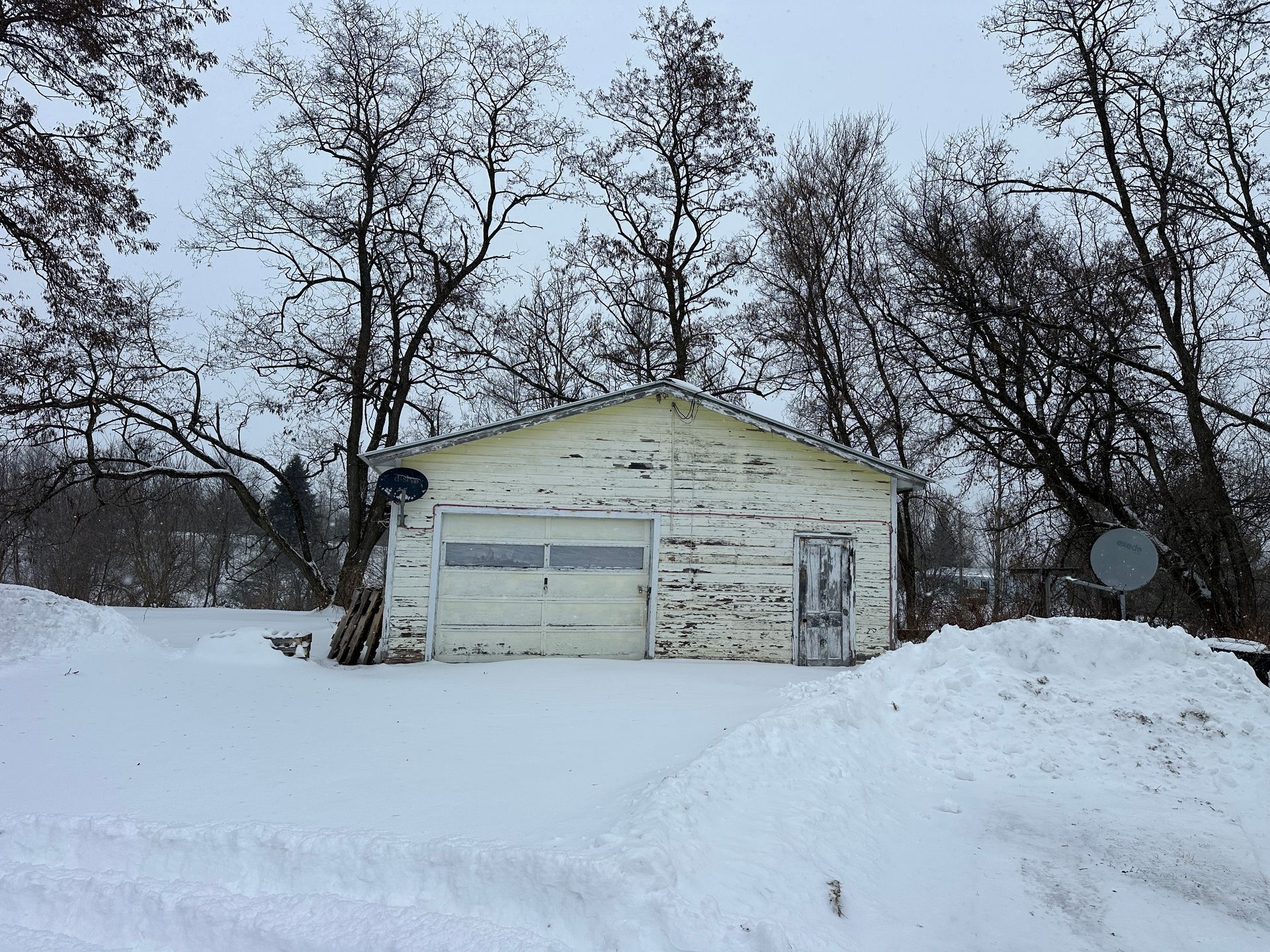 A white garage stands in a snow-covered field surrounded by bare trees under a gray winter sky.