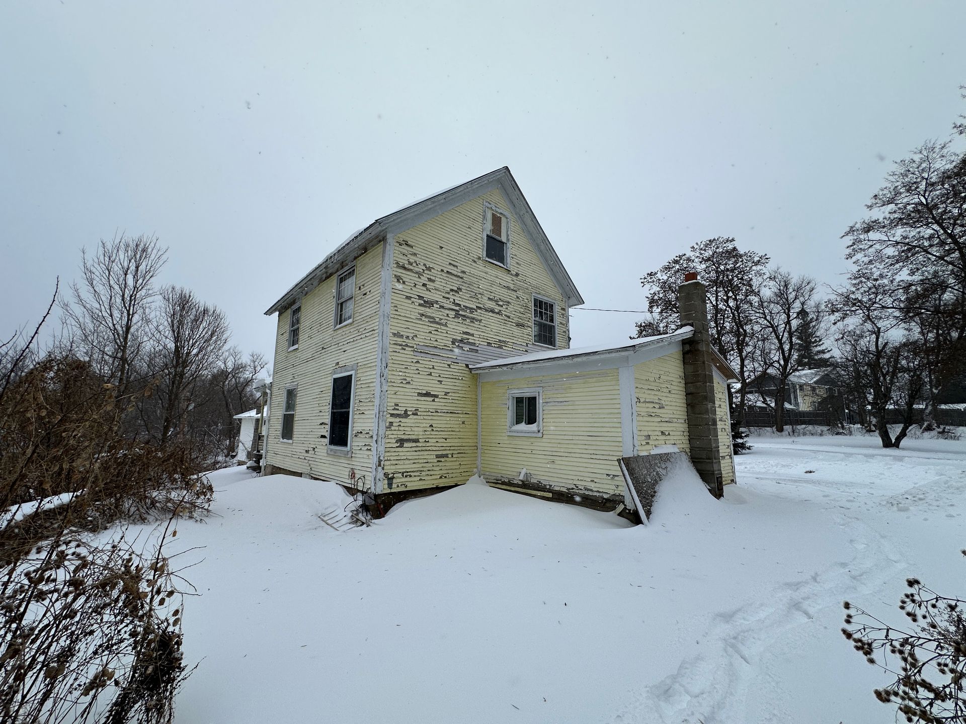 A yellow two-story house, surrounded by deep snow under a cloudy winter sky.