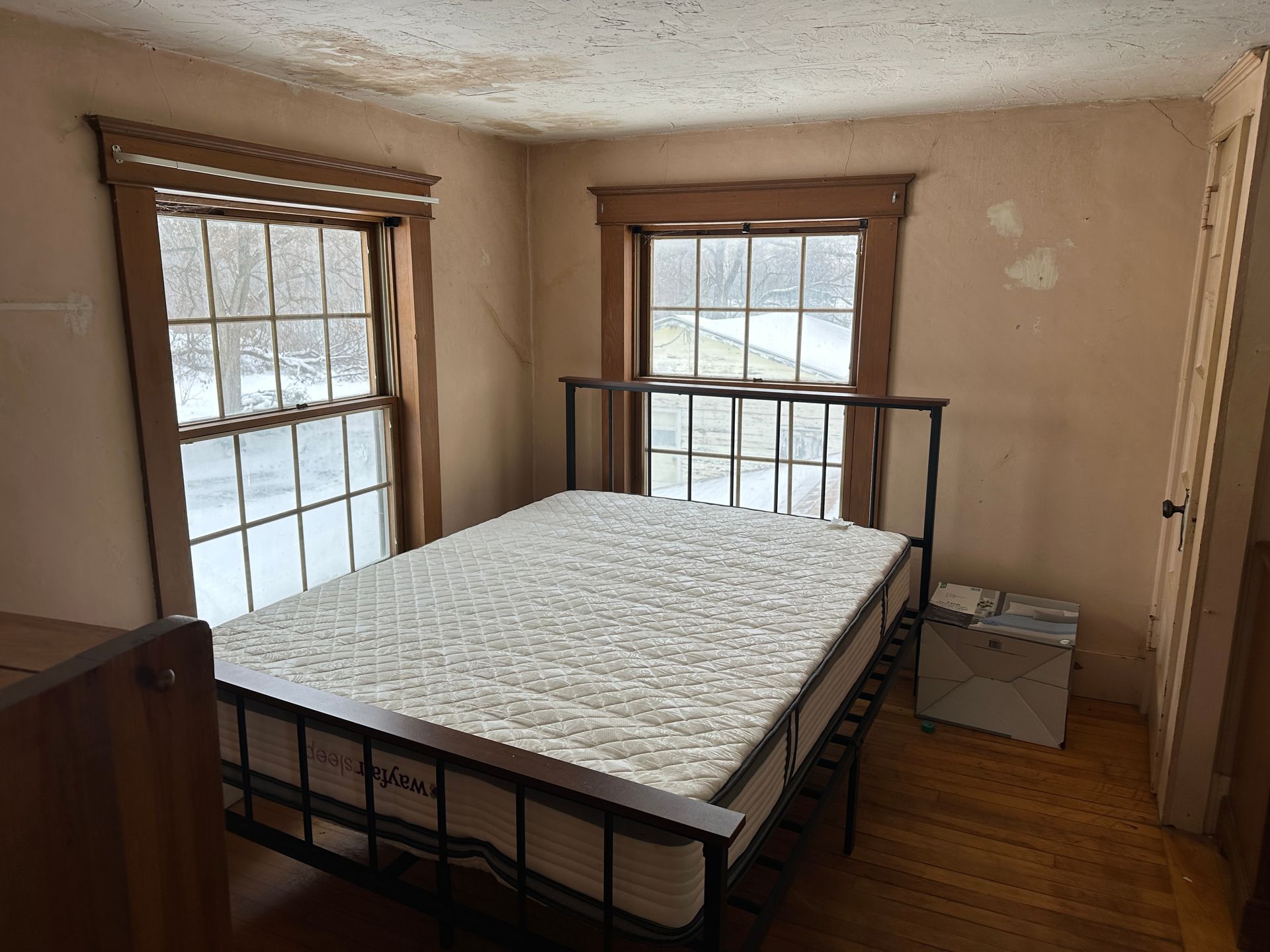 A sparsely furnished bedroom with a mattress on a black metal bed frame, two windows with wooden trim, and wood flooring.