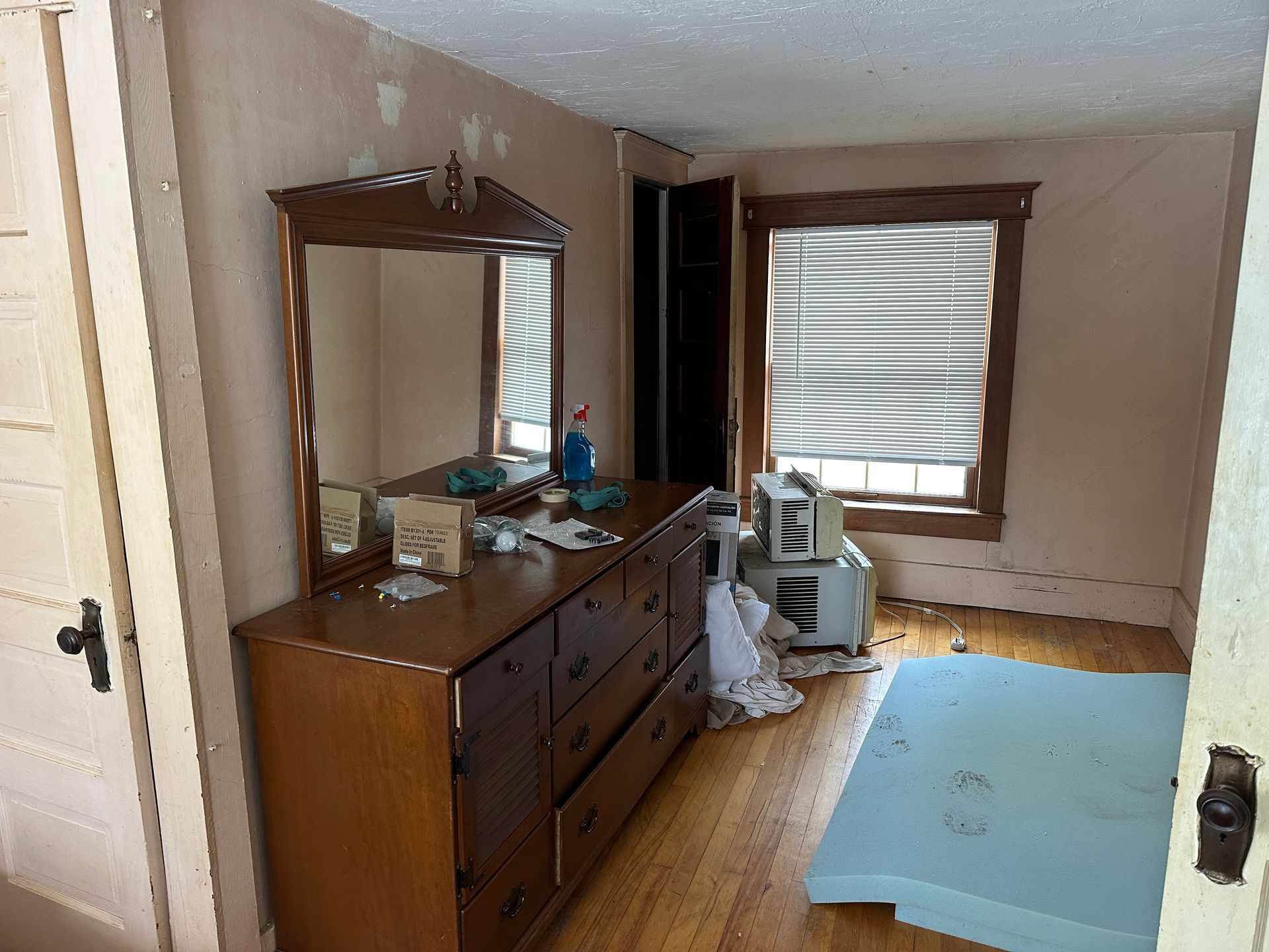 A bedroom featuring a wooden dresser with a mirror, a window with closed blinds, an air conditioner.