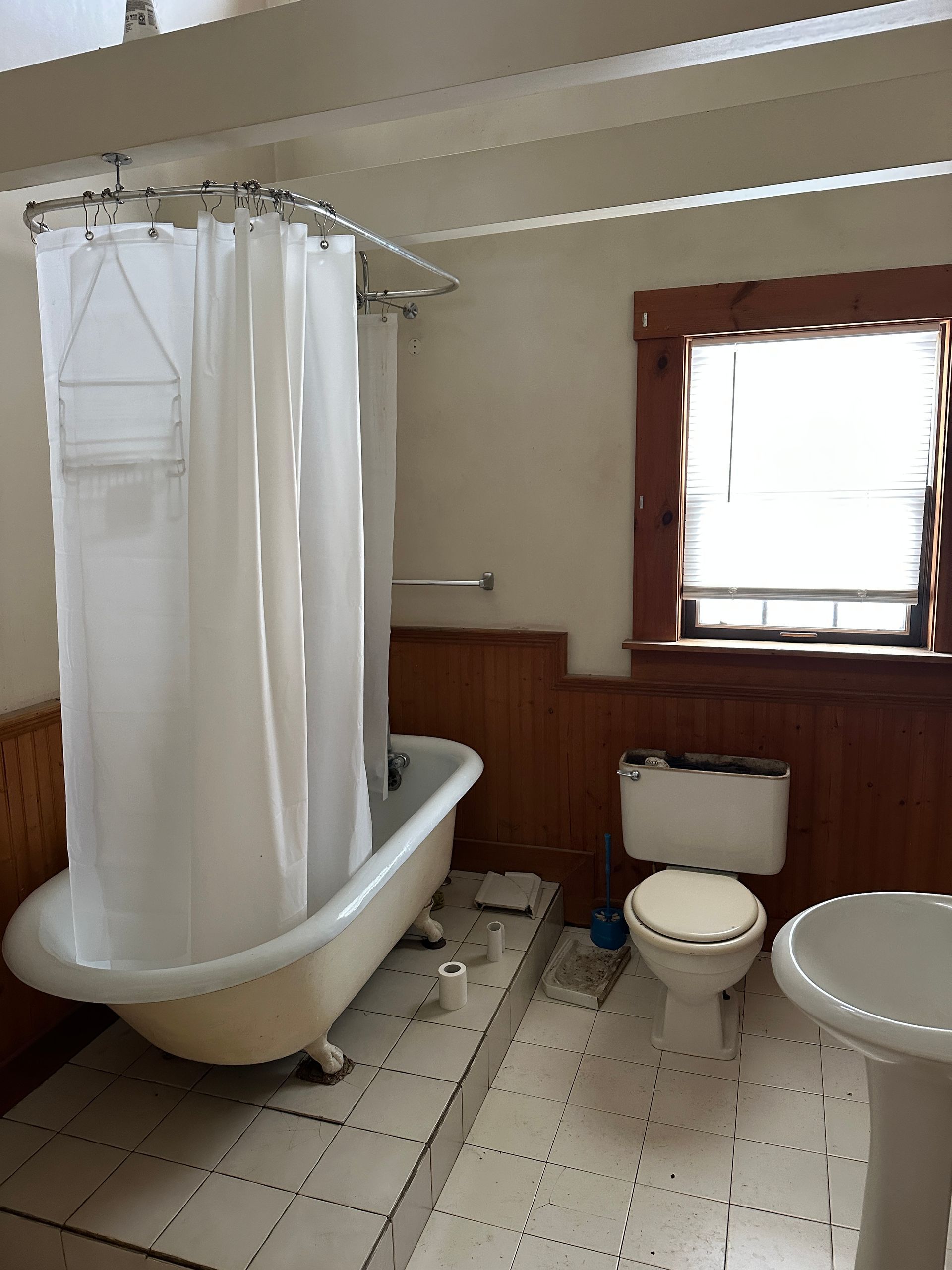 A vintage bathroom with a claw-foot tub, white shower curtain, toilet, and pedestal sink, featuring wood wainscoting.