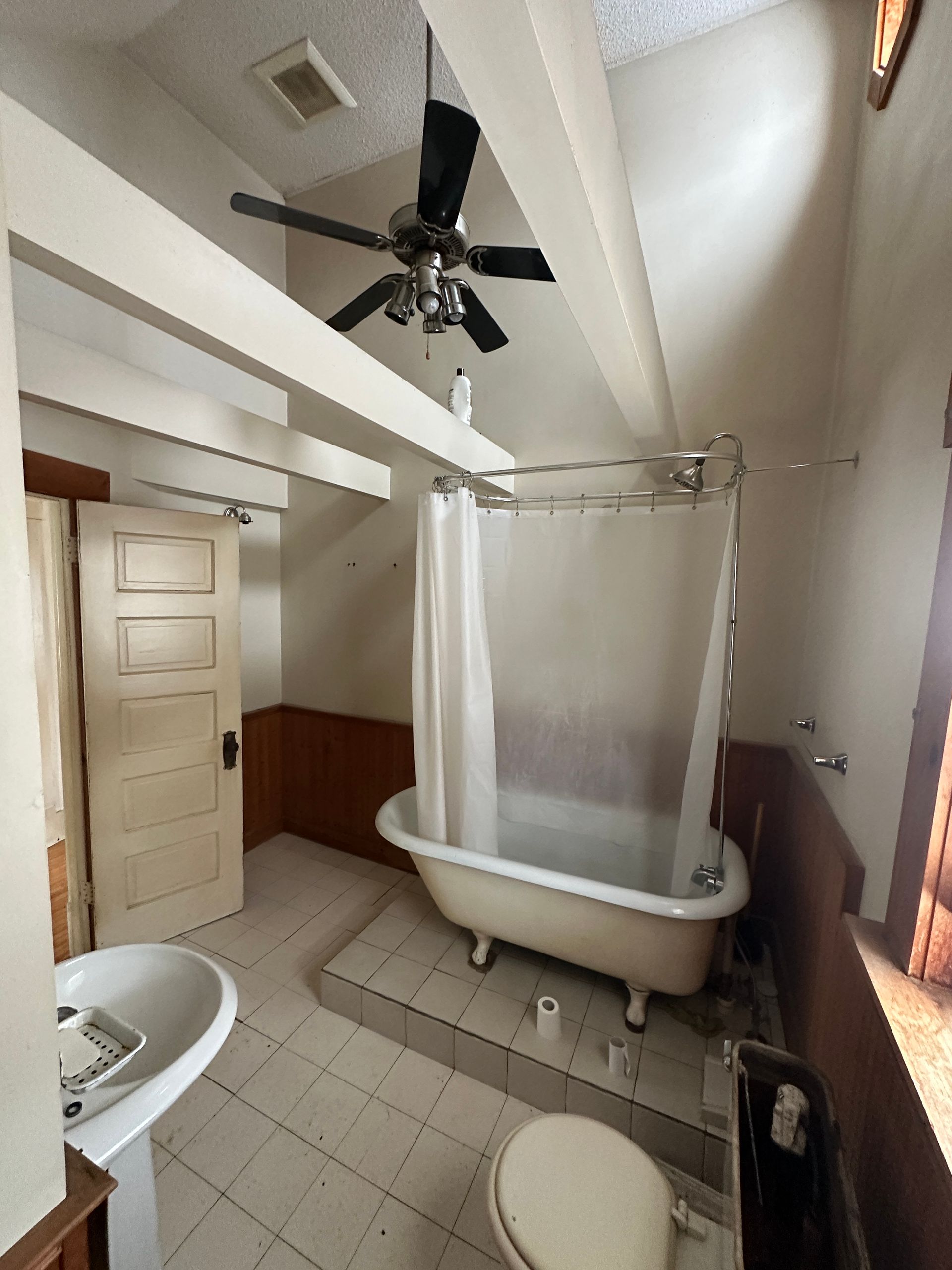 Bathroom with a claw-foot tub on a raised platform, pedestal sink, toilet, ceiling fan, and brown wall paneling.