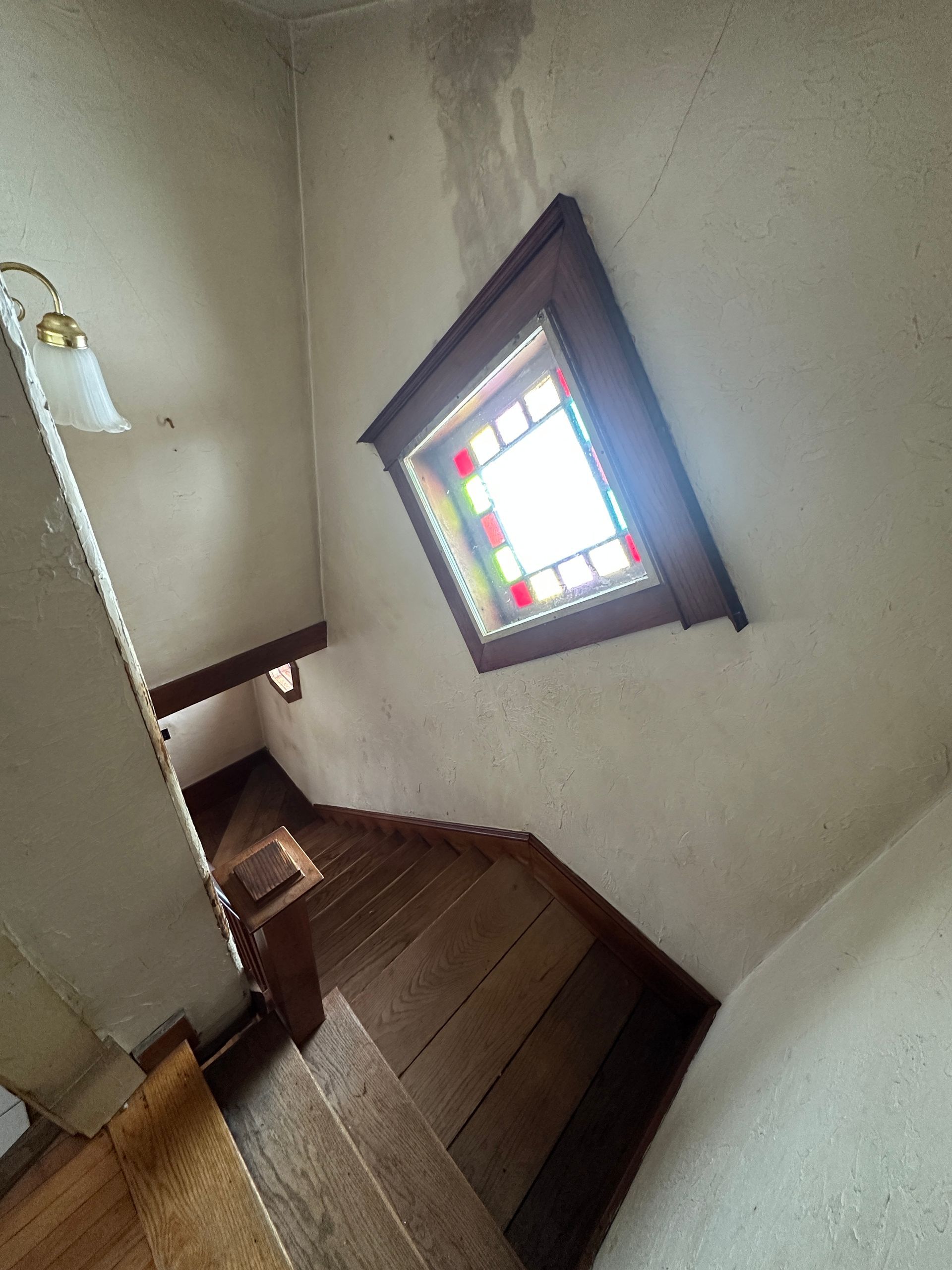 A wooden staircase leads downward toward a stained-glass window set into a plain wall.