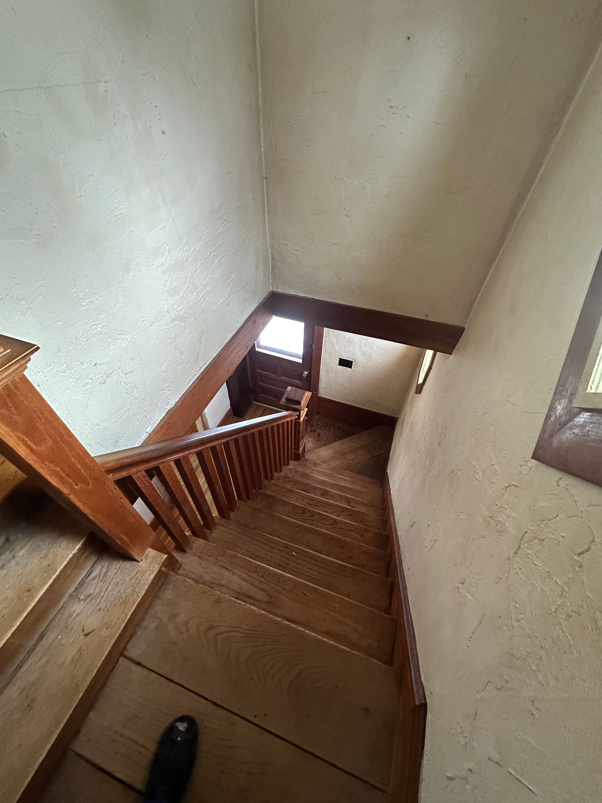 A high-angle view looking down a narrow wooden staircase toward a doorway with light shining through.